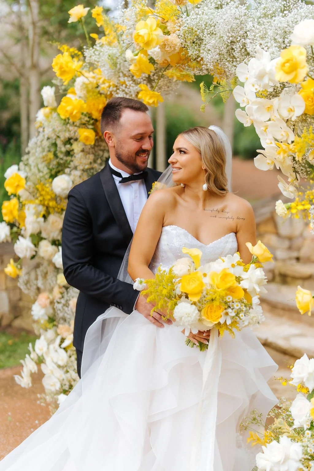 A bride and groom standing under a large floral arch with yellow and white flowers, smiling at each other during their wedding ceremony outdoors.