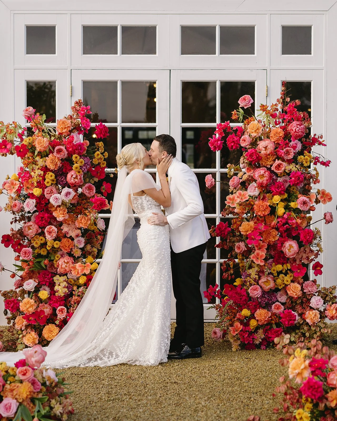 A bride and groom sharing a kiss in front of a large floral arch on their wedding day.