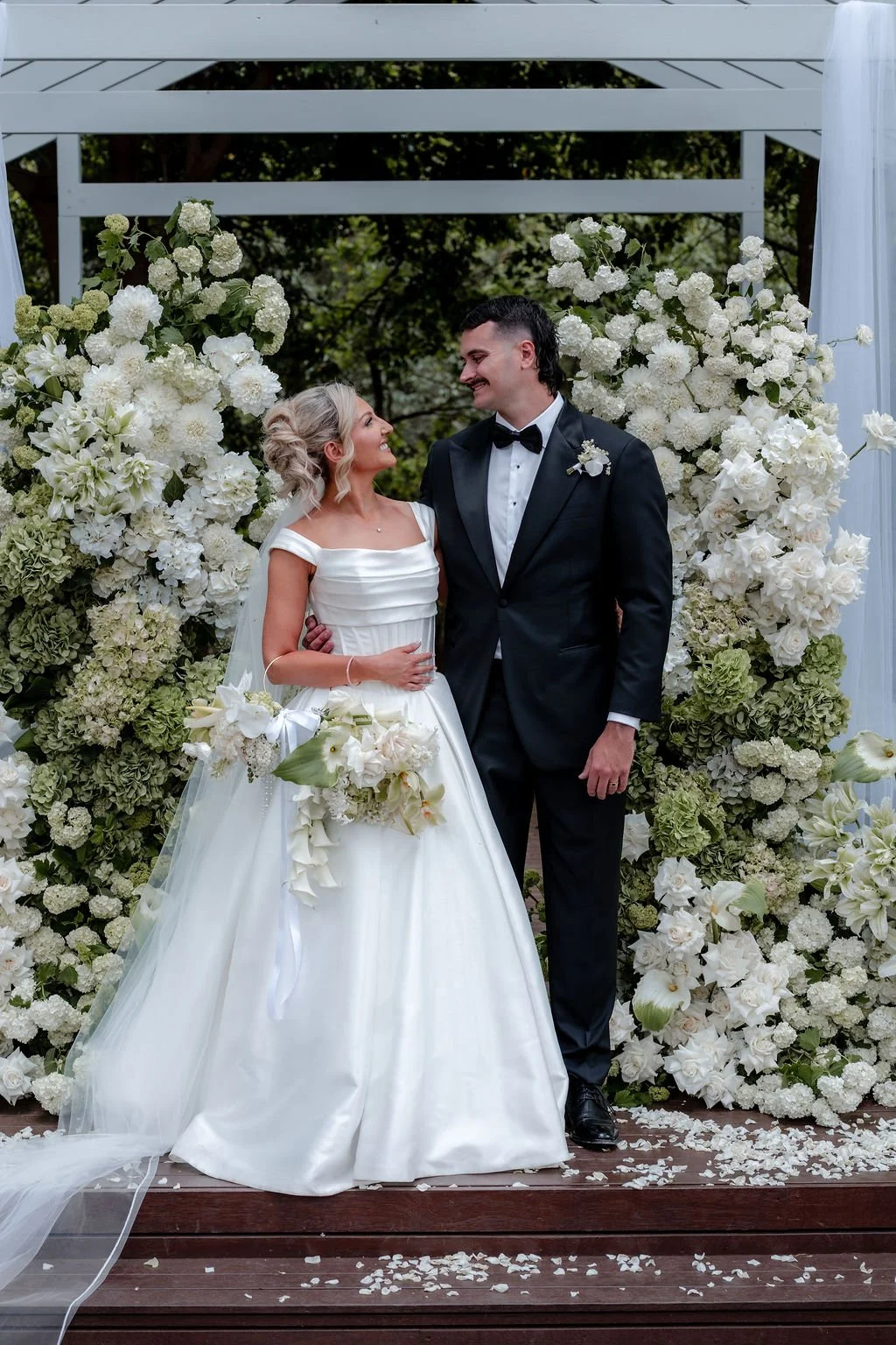A bride and groom standing together on their wedding day, surrounded by white floral arrangements, outdoors beneath a white arch.