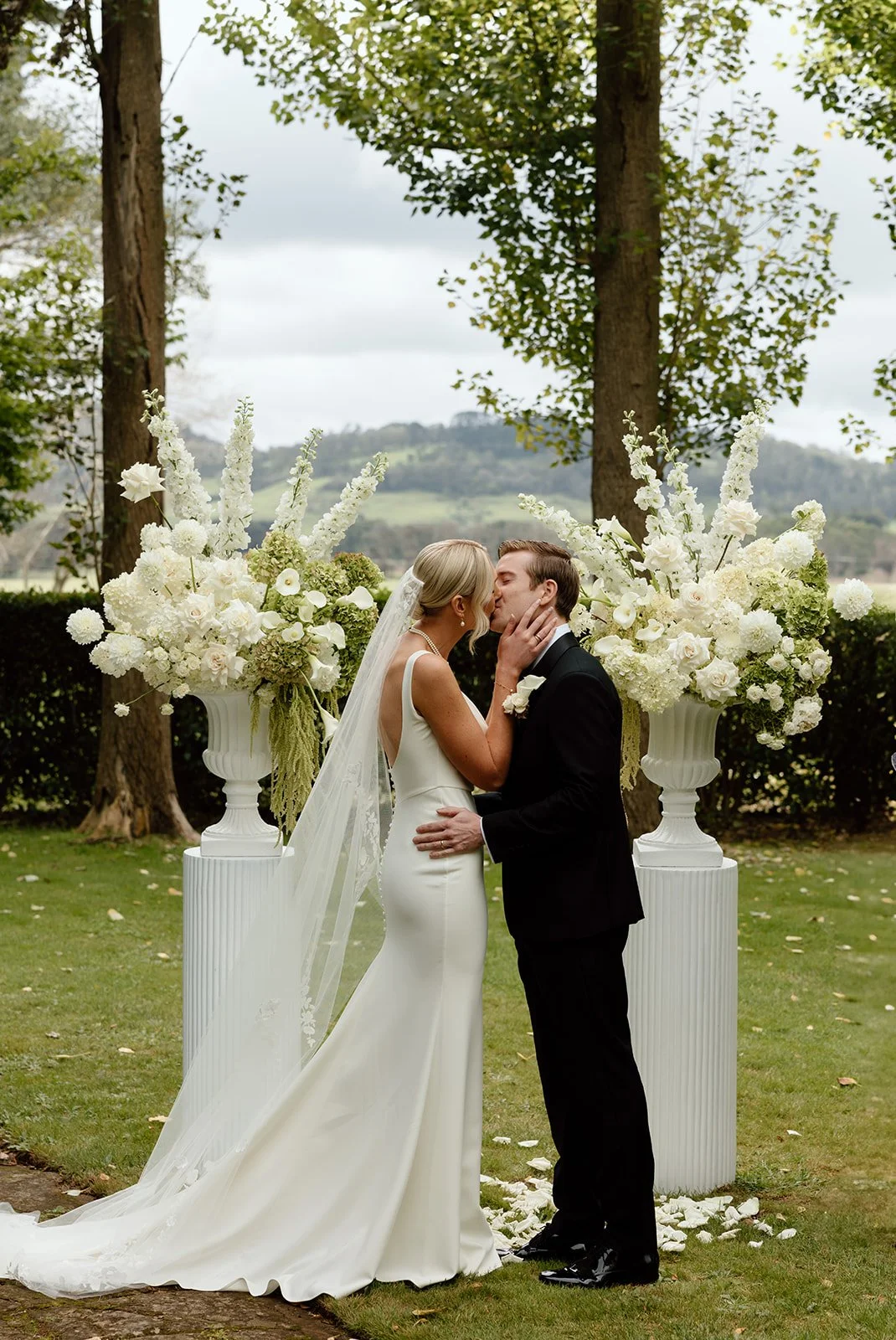 A bride and groom share a kiss at their outdoor wedding ceremony, standing in front of large floral arrangements of white flowers and greenery, with trees and hills in the background.