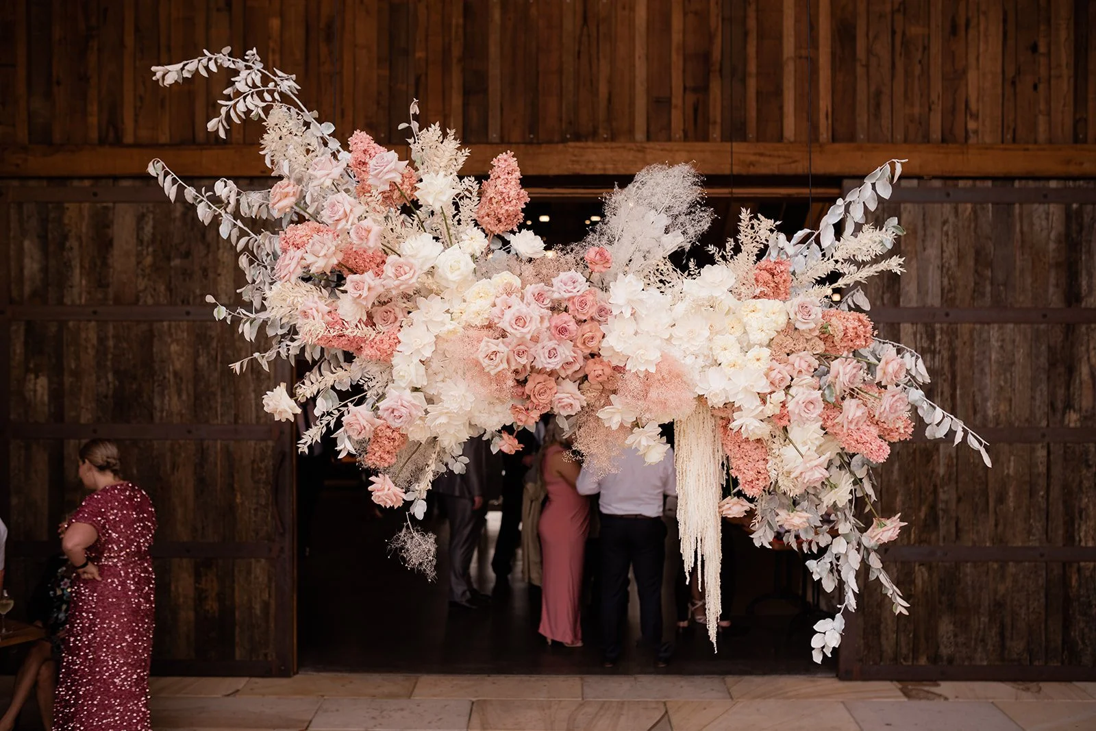Large decorative floral arrangement with pink, white, and peach flowers and white foliage hanging above a wooden doorway at an indoor event.