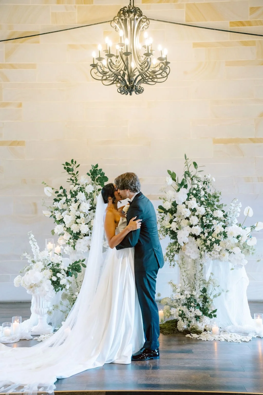 Kissing bride and groom at wedding ceremony with floral arrangements and candles.