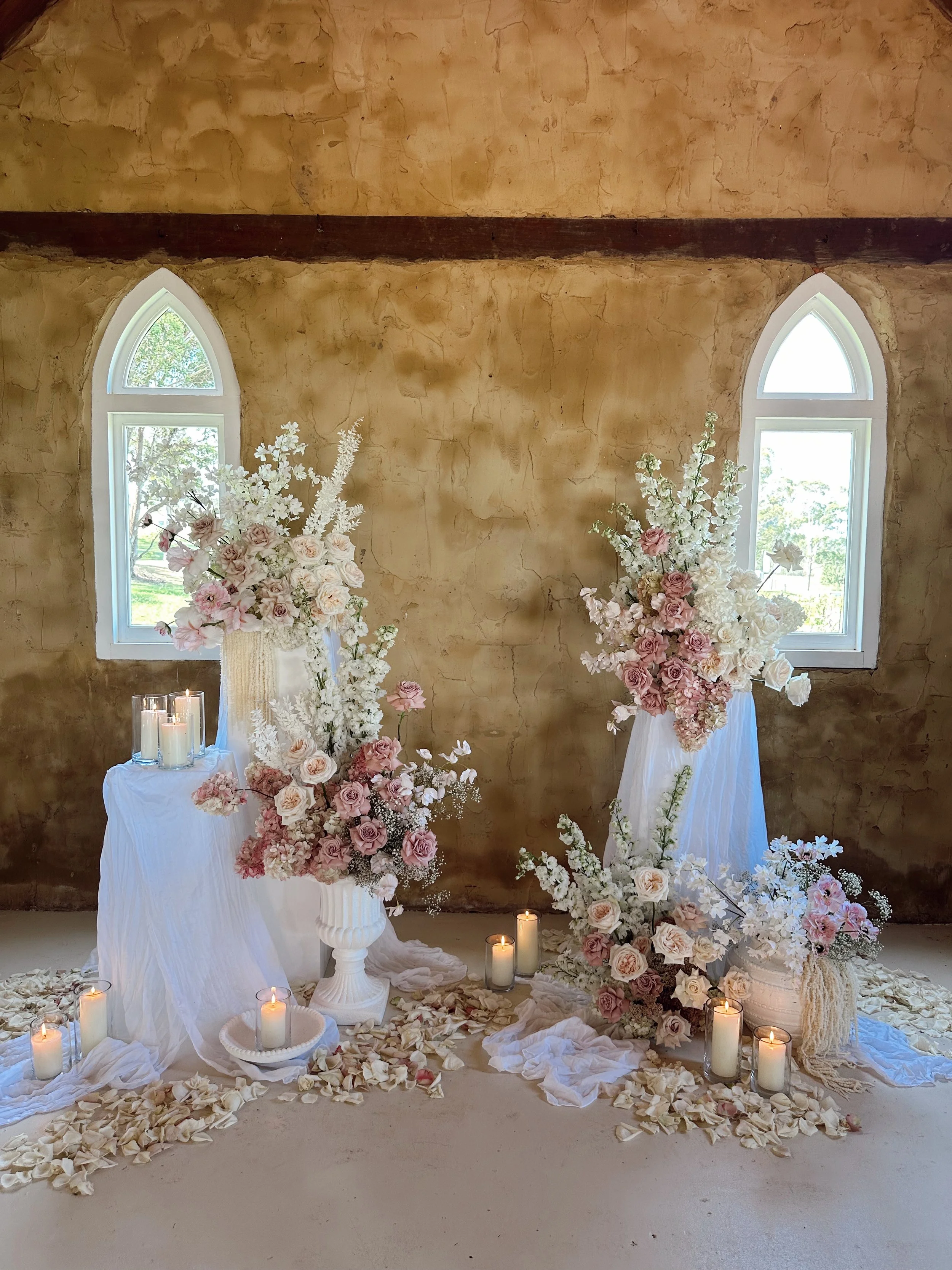 A romantic floral arrangement with pink and white roses, peonies, and white hydrangeas in tall vases, surrounded by white candles and flower petals, set against a rustic wall with two arched windows.