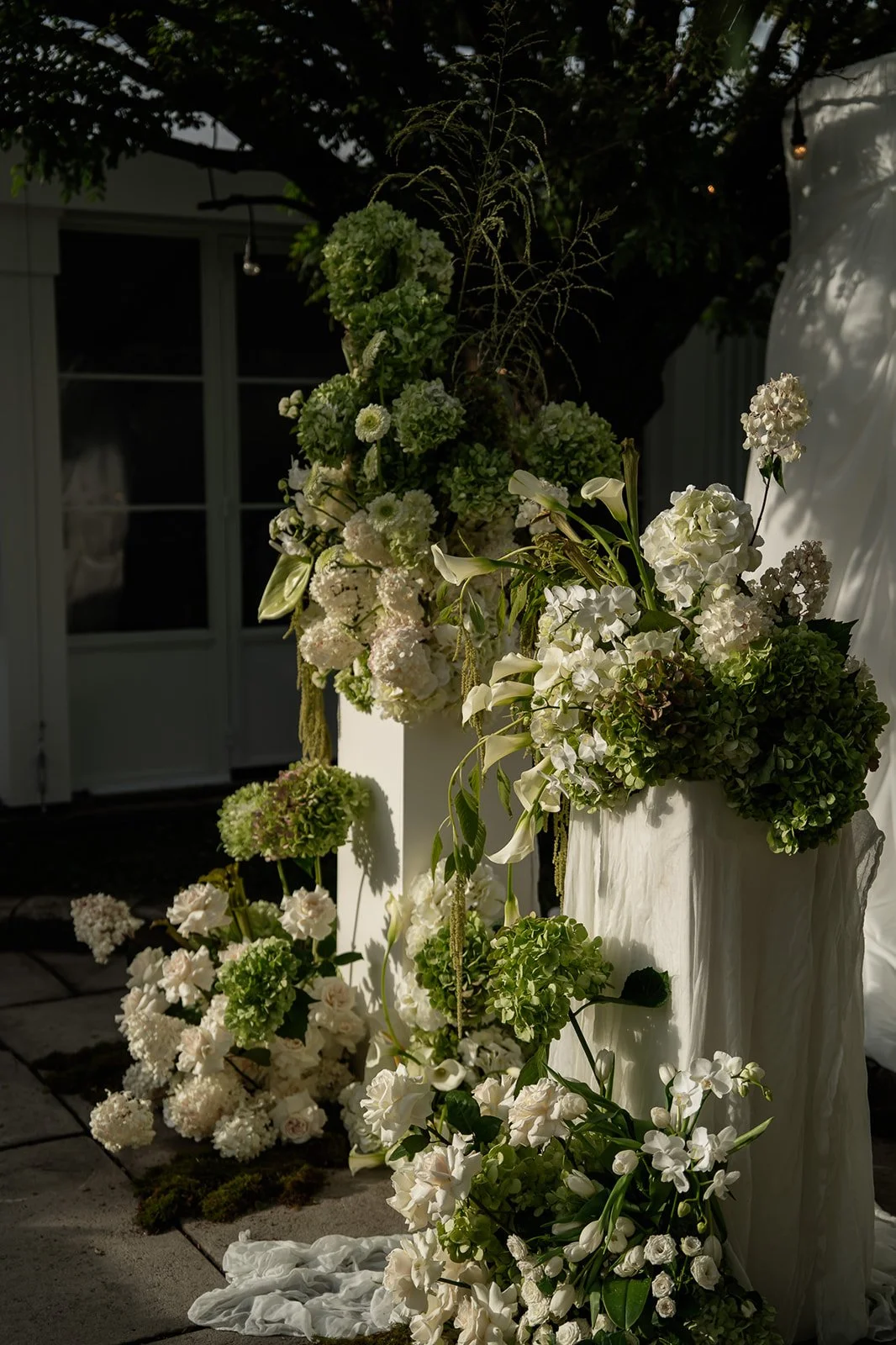 Bouquet of white and green flowers, including hydrangeas, calla lilies, and lisianthus, arranged on a white fabric-covered stand outdoors with sunlight and shadow.