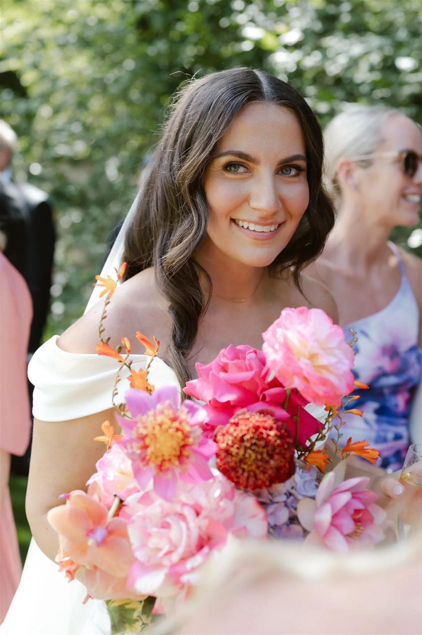 A bride holding a colorful bouquet of pink, orange, and red flowers at an outdoor wedding.