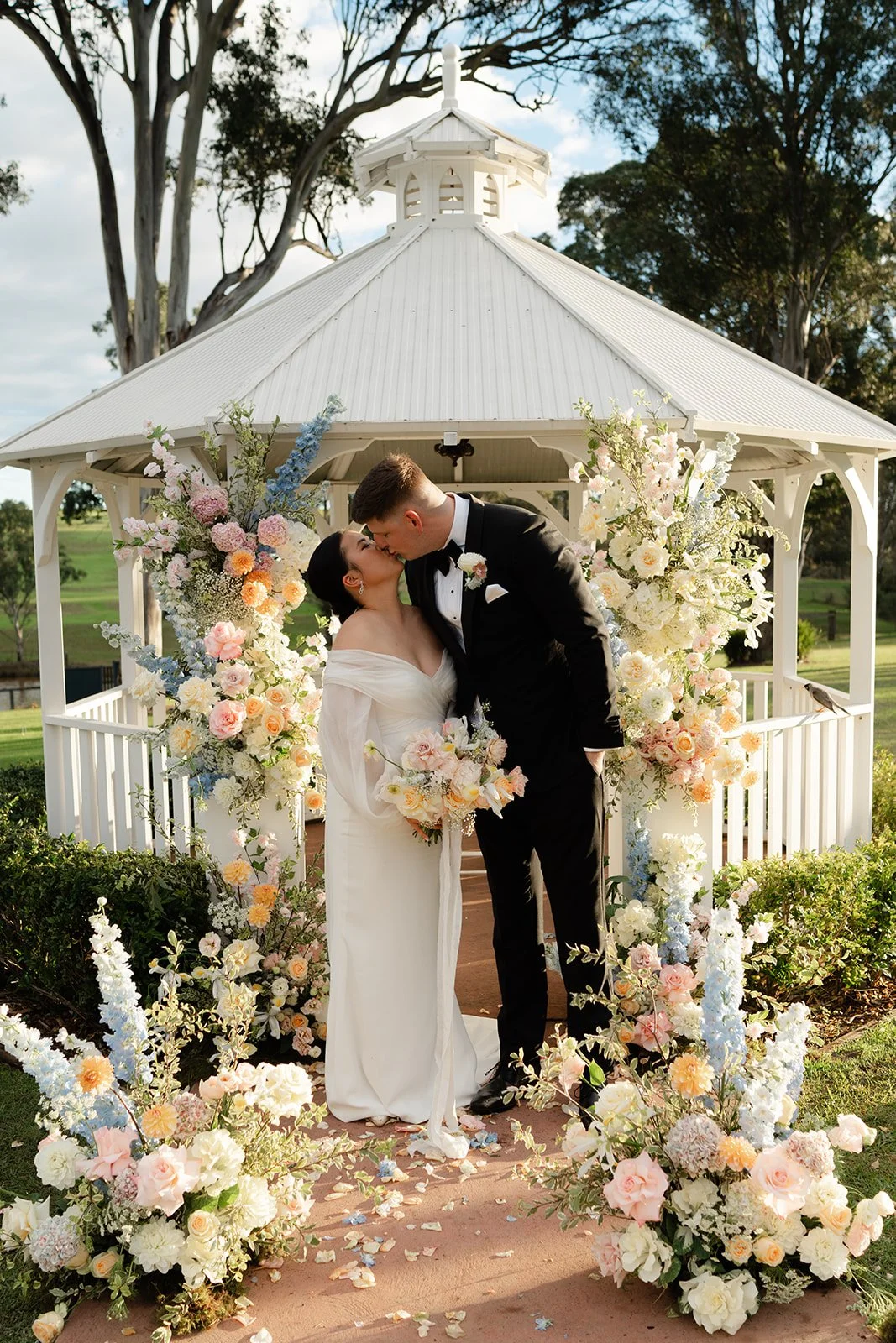 A bride and groom kissing under a white gazebo decorated with pink, white, and blue flowers during their wedding.