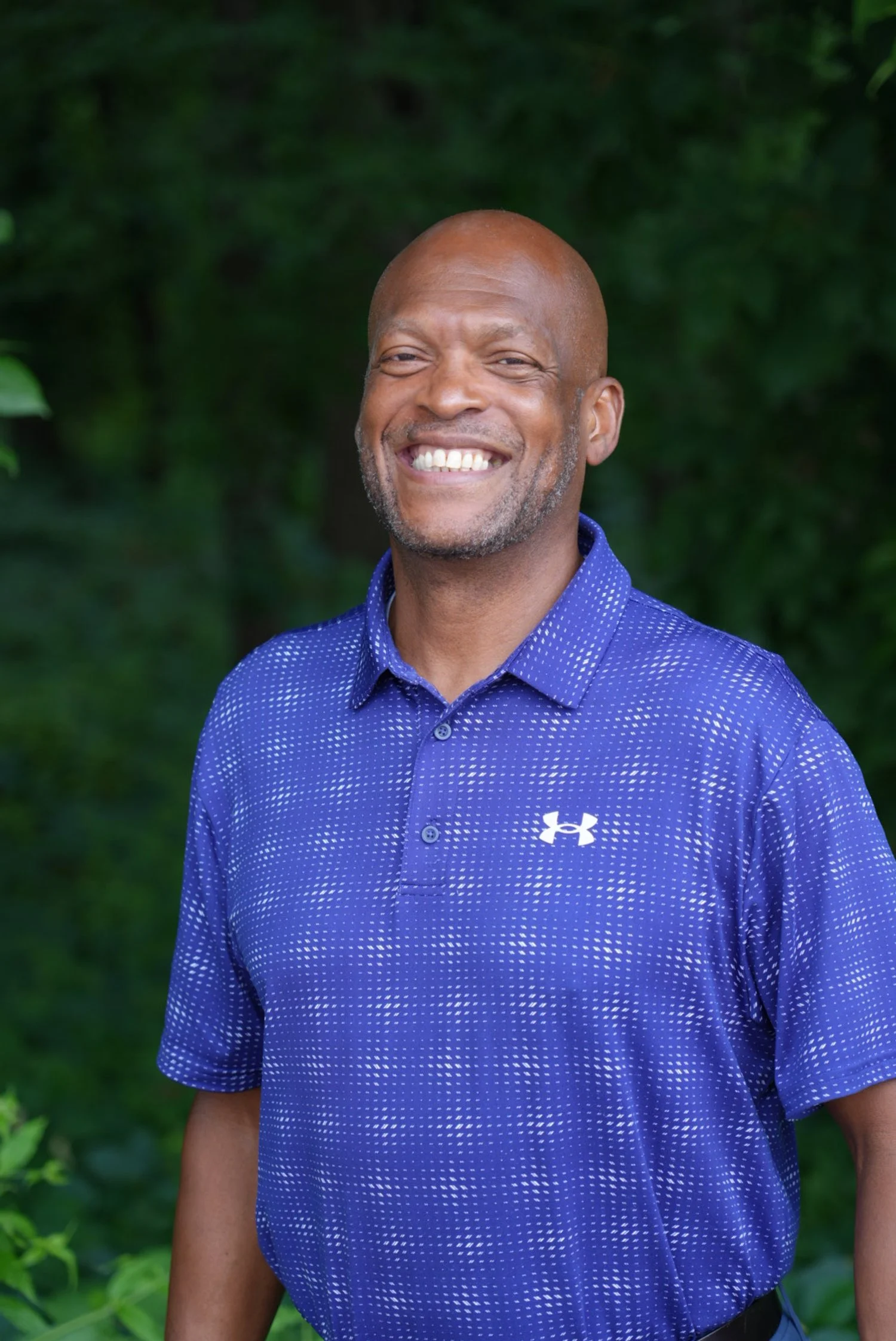 A smiling man wearing a blue golf shirt, standing against green foliage