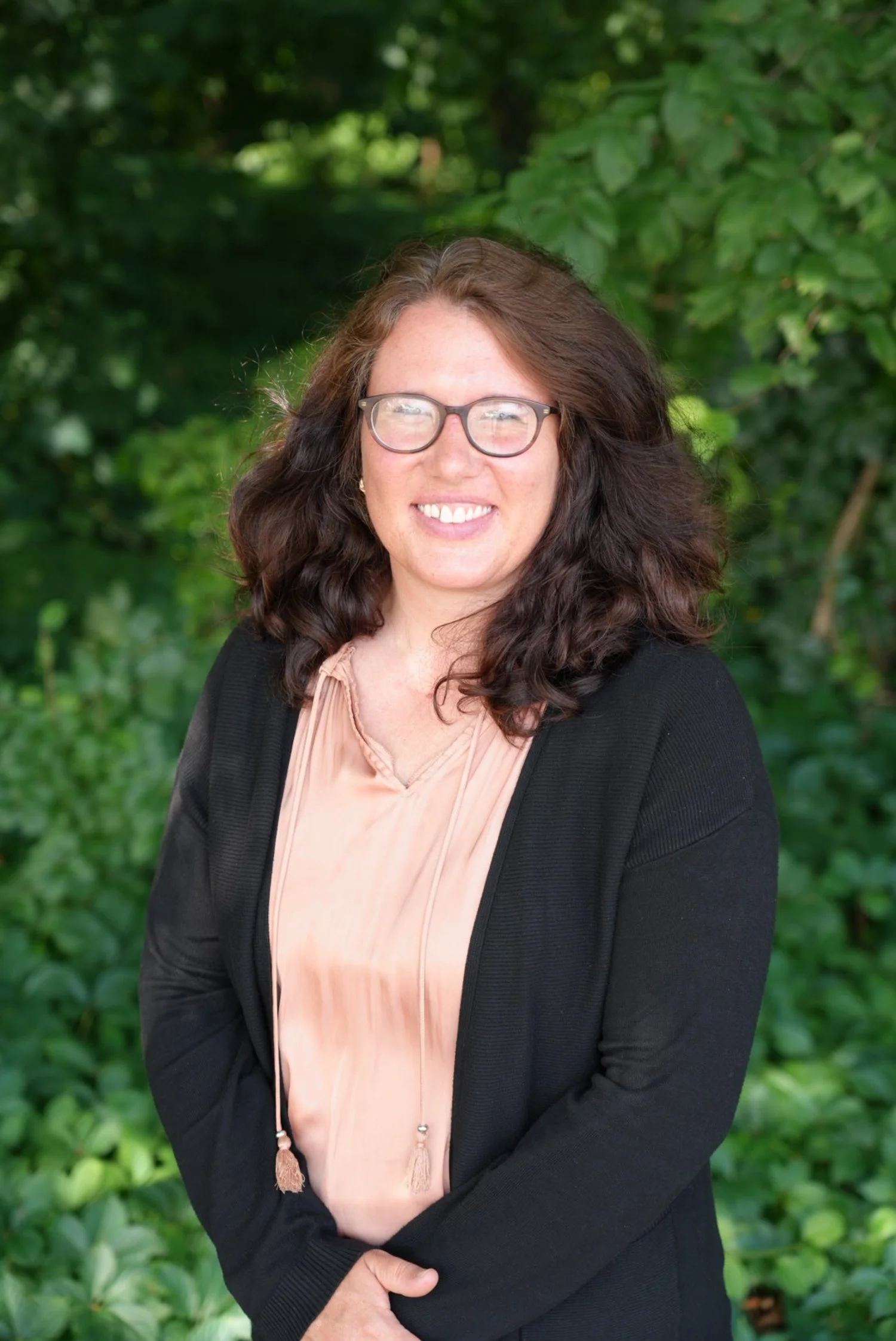 A woman with long, wavy brown hair and glasses, smiling, wearing a dark gray turtleneck sweater, standing against a plain light-colored wall.