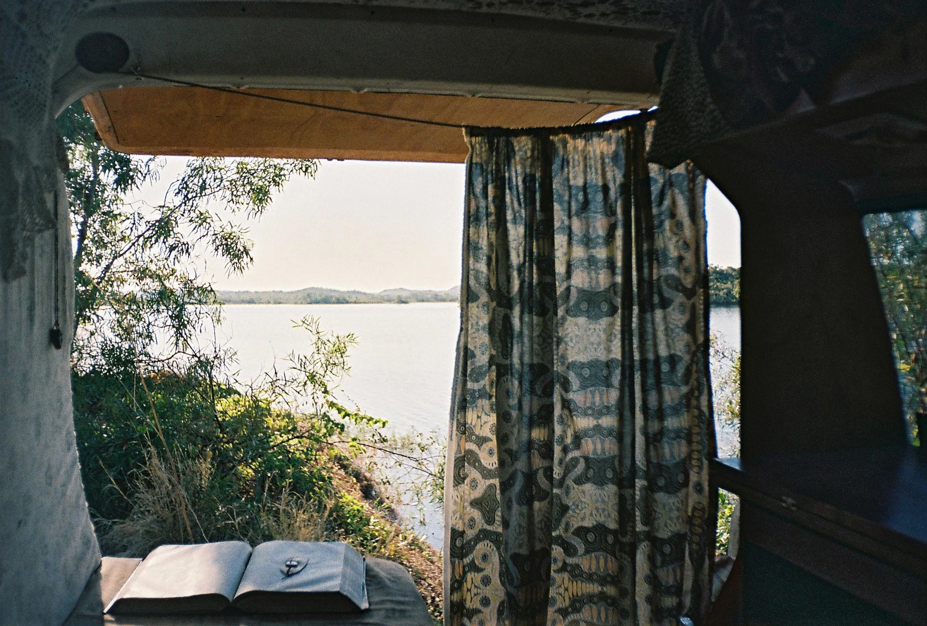 Photo from inside a caravan looking out at a blue curtain, a little desk with a bible open on it and a river in front of it