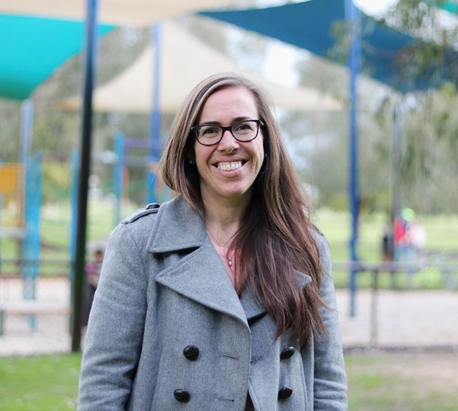 Photo of the author in a gray coat and pink shirt in front of a playground