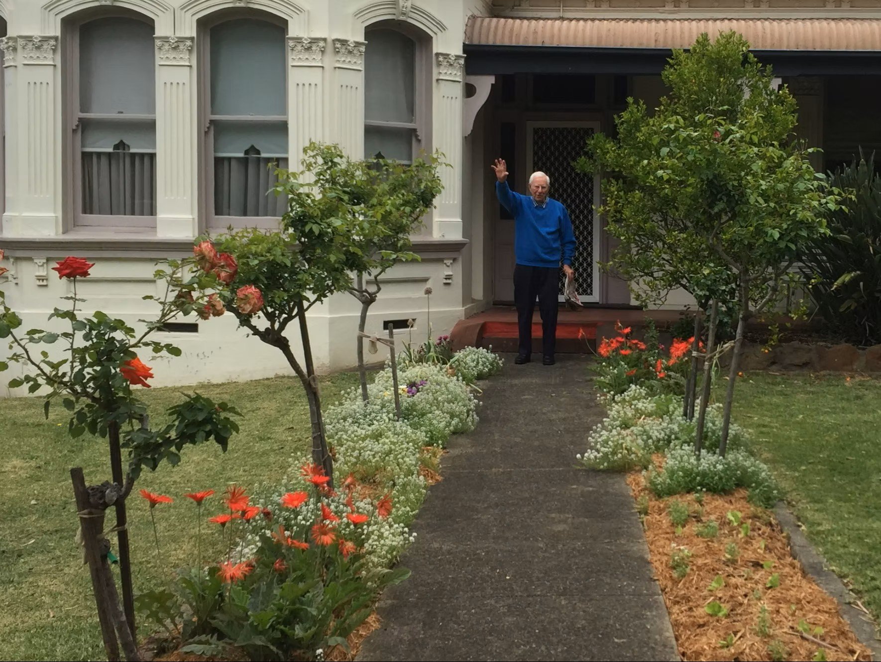 Photo of the author's grandfather wearing a blue knit jumper and slacks, waving out the front of his home with roses and garden