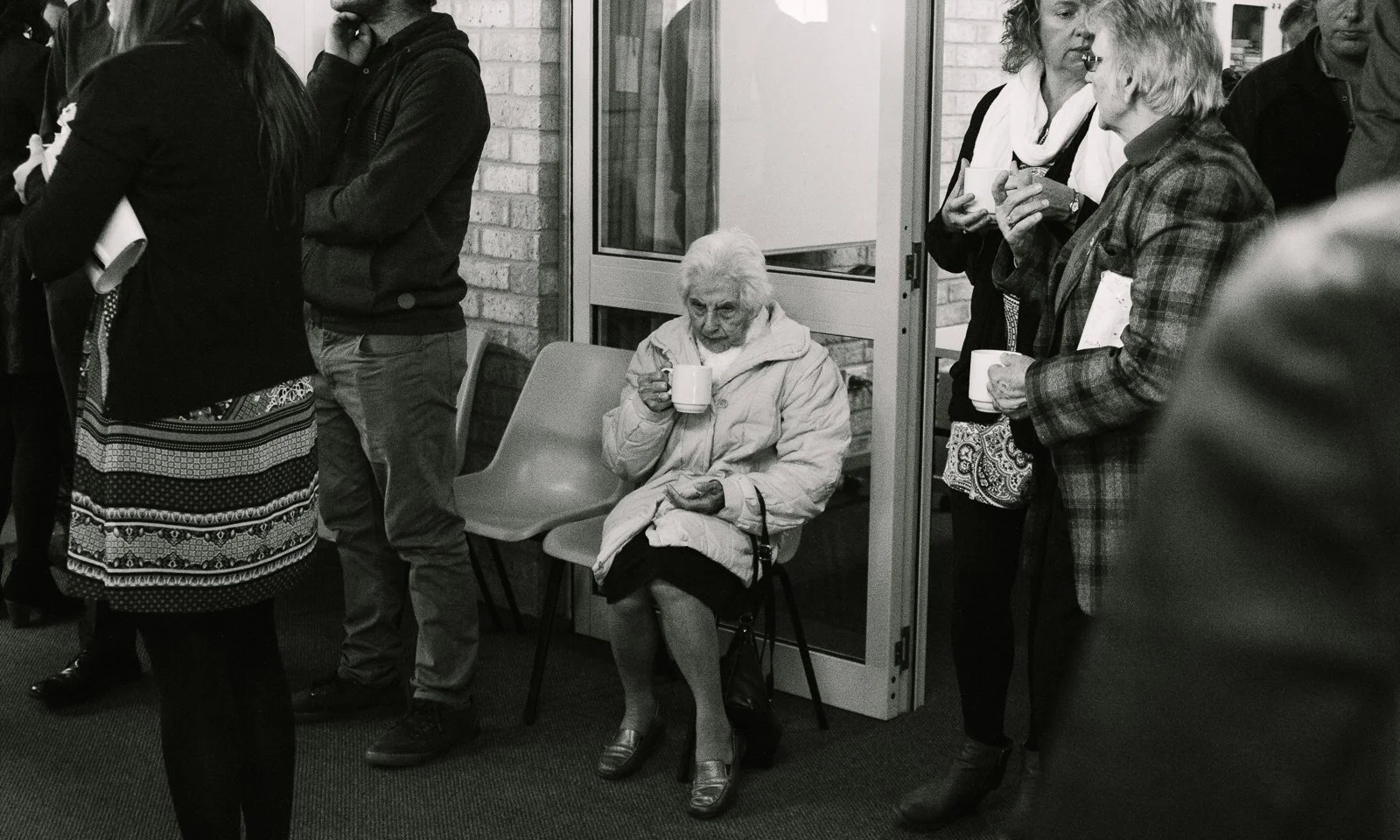 old woman sitting down and drinking a cup of tea while surrounded by people