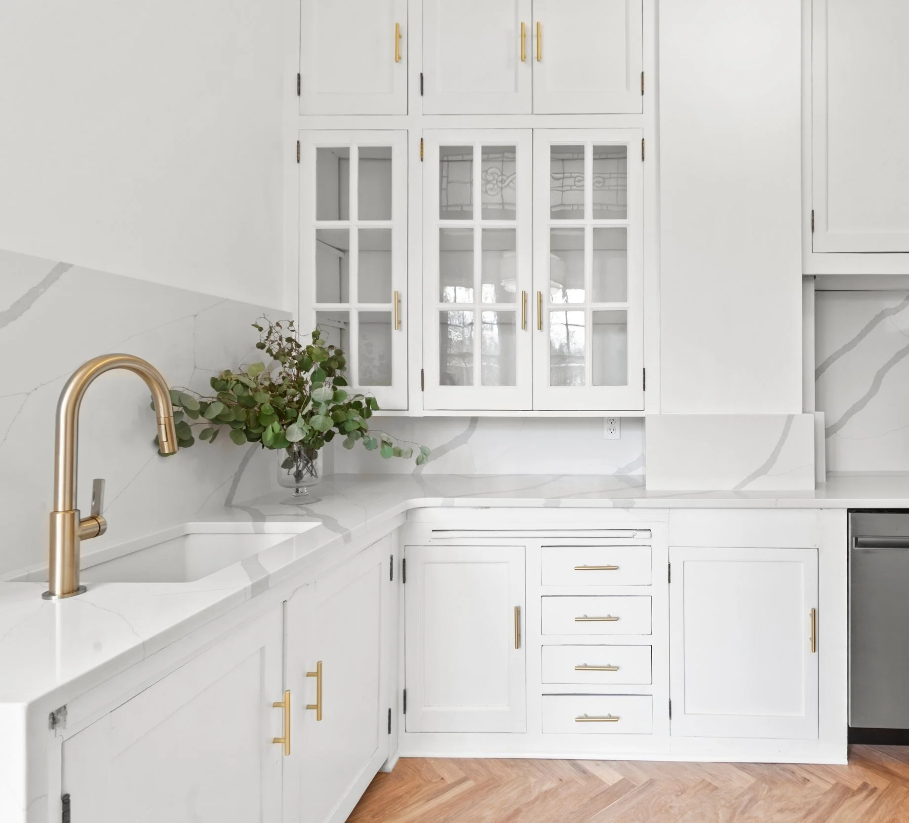 Modern white kitchen with gold faucet, marble countertops, glass-front cabinets, and herringbone wood flooring.