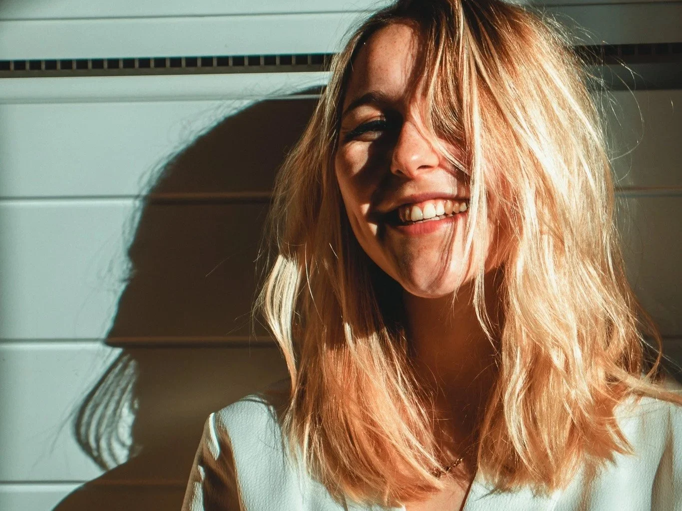 Lächelnde junge Frau mit lockigem, blondem Haar, die im Sonnenlicht steht und neben einer weißen Wand sitzt.
