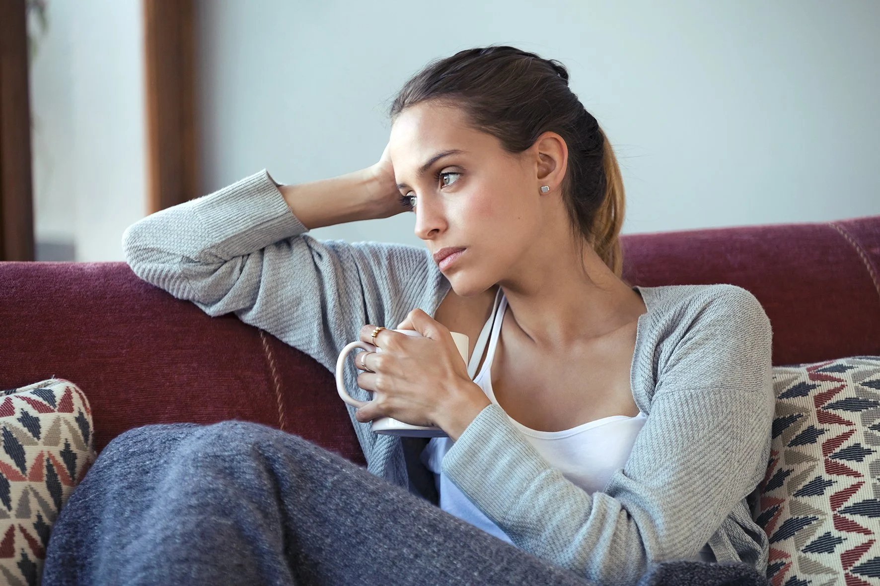 Eine junge Frau sitzt auf einem roten Sofa, hält eine weiße Tasse in der Hand und wirkt nachdenklich. Sie trägt eine graue Strickjacke und ein weißes Top, mit braunen Haaren, die zu einem Pferdeschwanz gebunden sind.