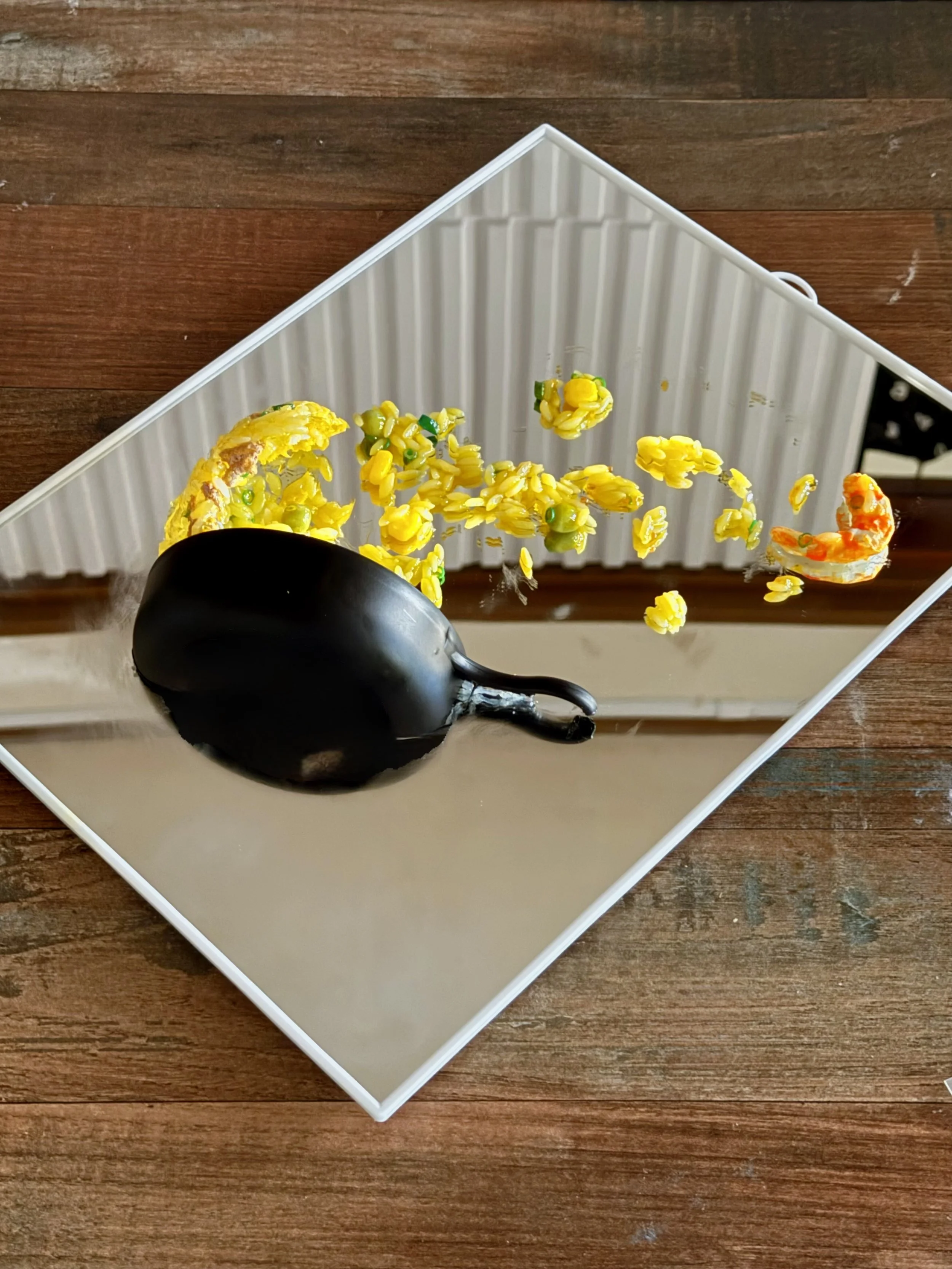 A black fried pan tipped over on a white tray with yellow fried rice and a small shrimp. The tray is on a mirror.
