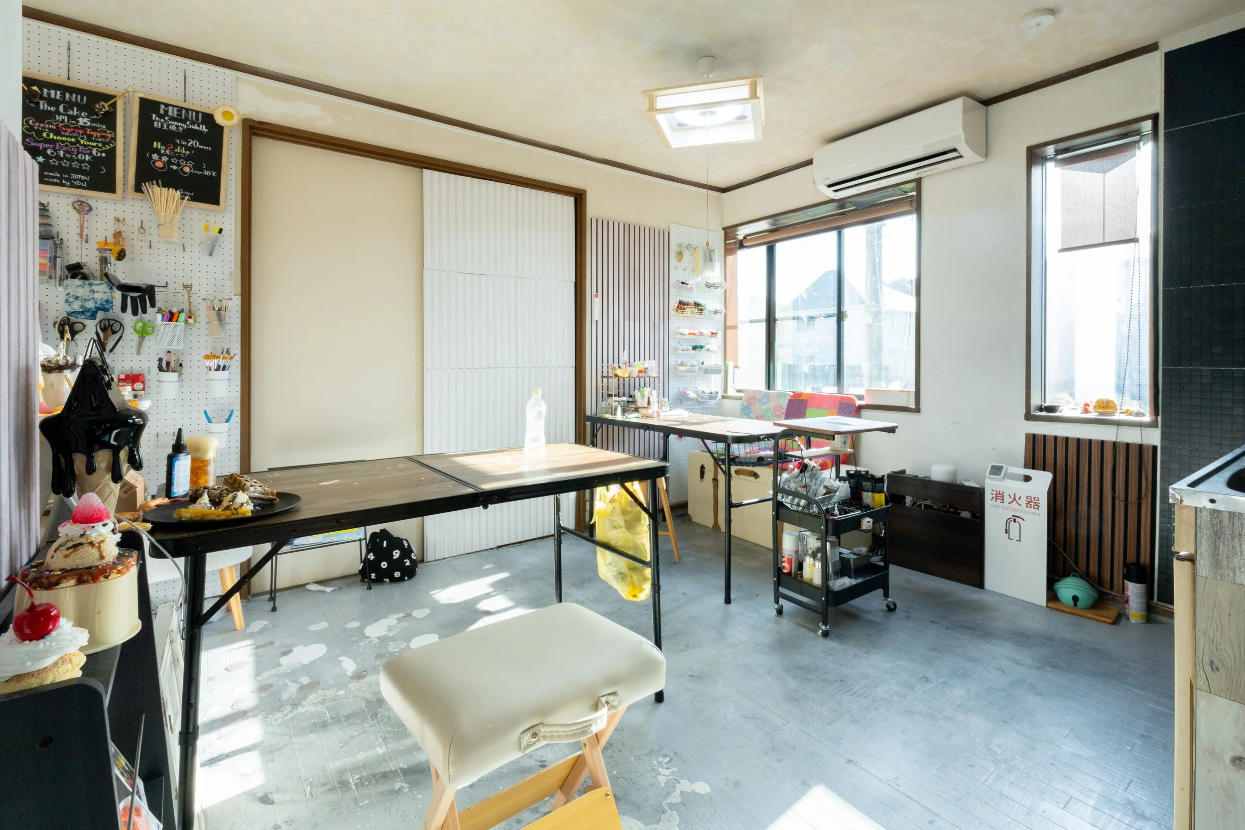 Interior of a workspace with tables, chairs, and various supplies, including a black pegboard with tools and utensils, large windows letting in natural light, and a collapsed table with spilled paint or plaster on the floor.