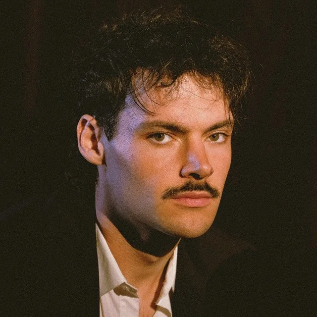 A young man with dark, curly hair, a mustache, wearing a black blazer and white shirt, looking directly at the camera against a dark background.