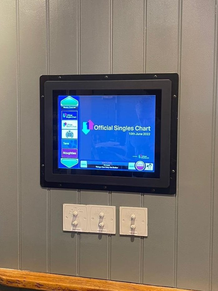 A wall-mounted digital screen displaying the Official Singles Chart dated 10th June 2022, with music categories on the left side. Below the screen, there are multiple volume control knobs and sockets.