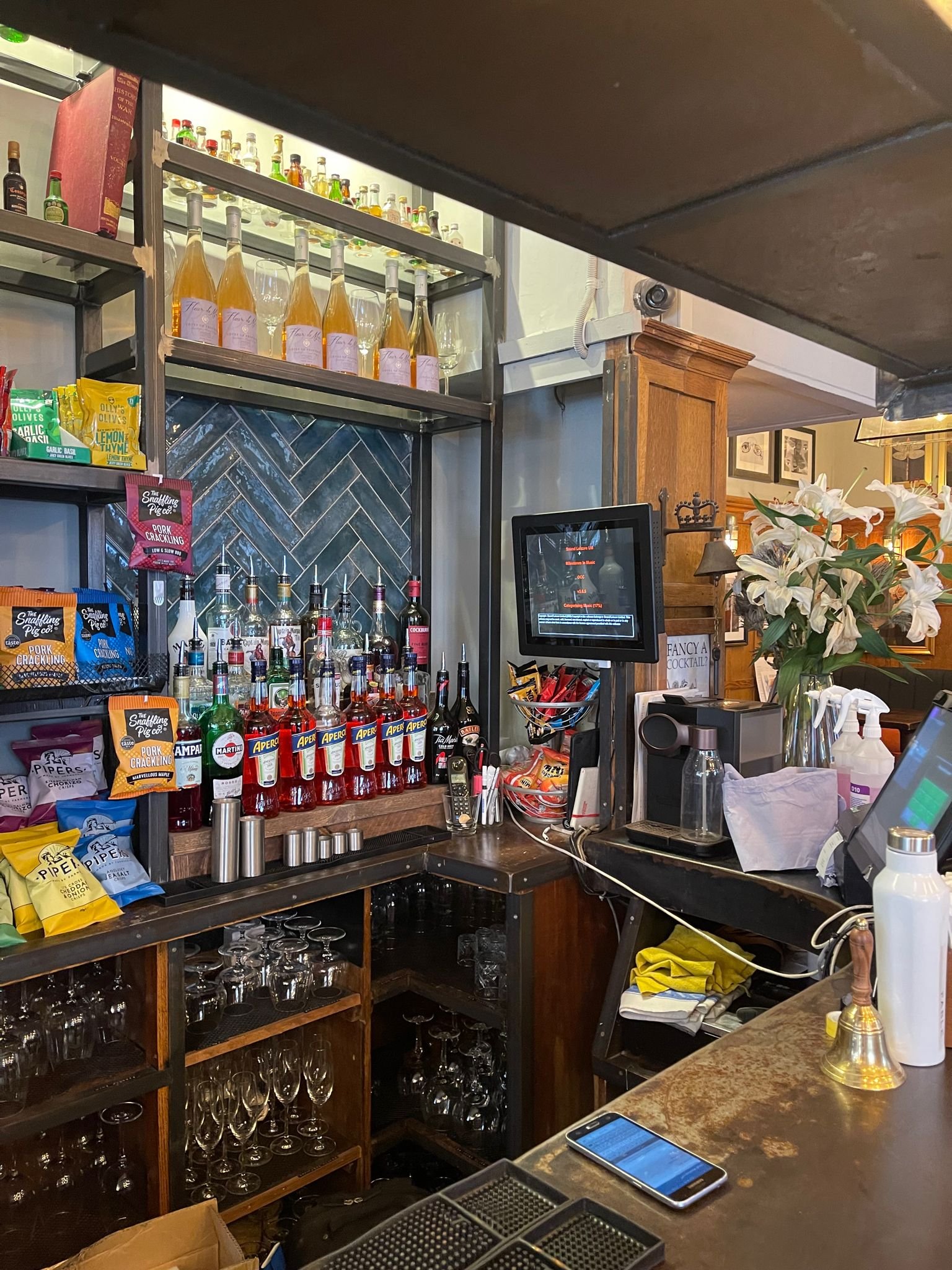 Interior of a bar with shelves of liquor bottles, snacks, and glassware behind a counter. A digital screen, flower vase, sanitizers, and a smartphone are seen on the counter.