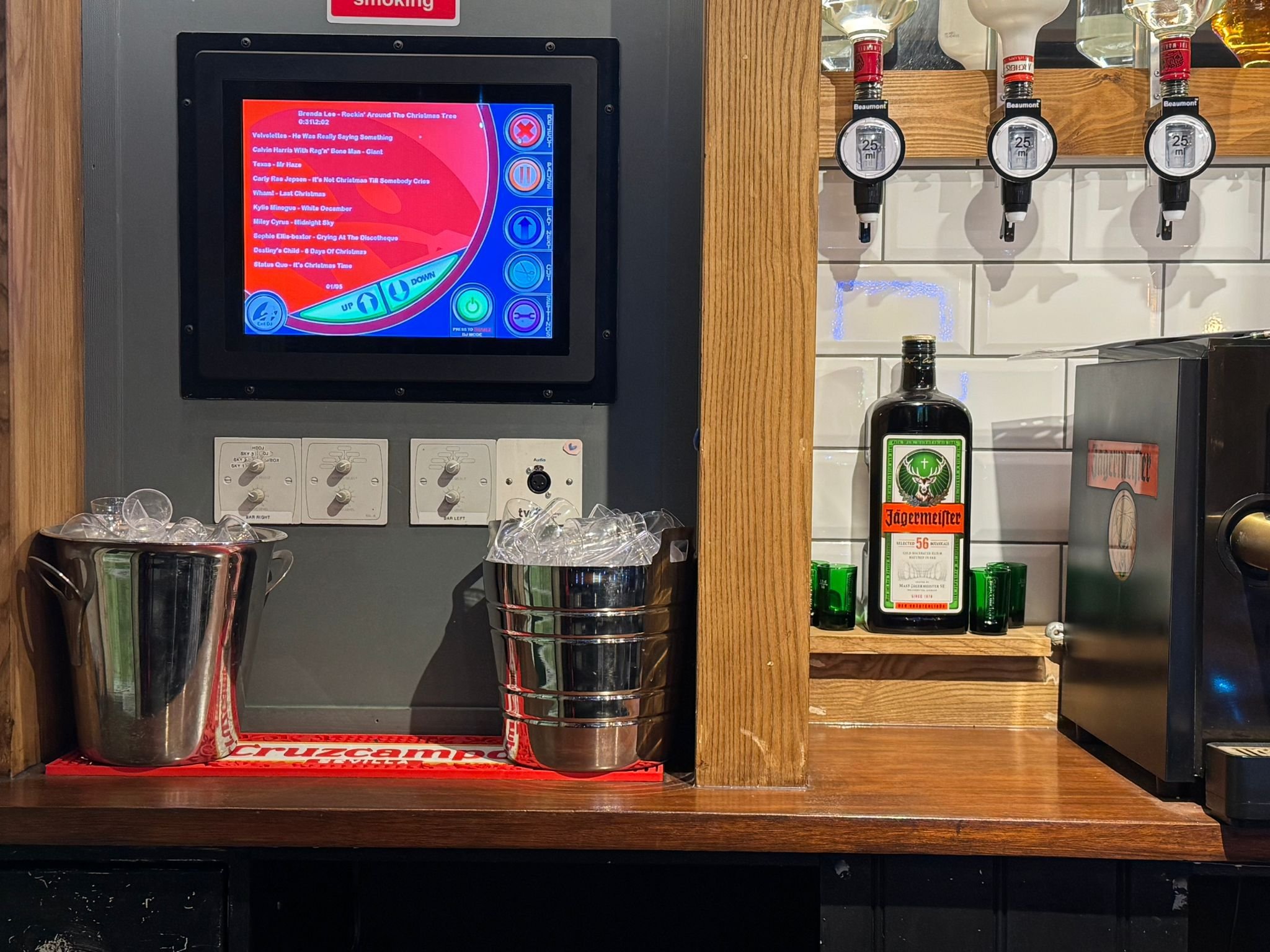 Bar setup with a touchscreen music or karaoke system, ice buckets, drink dispensers, bottles, and Jägermeister.