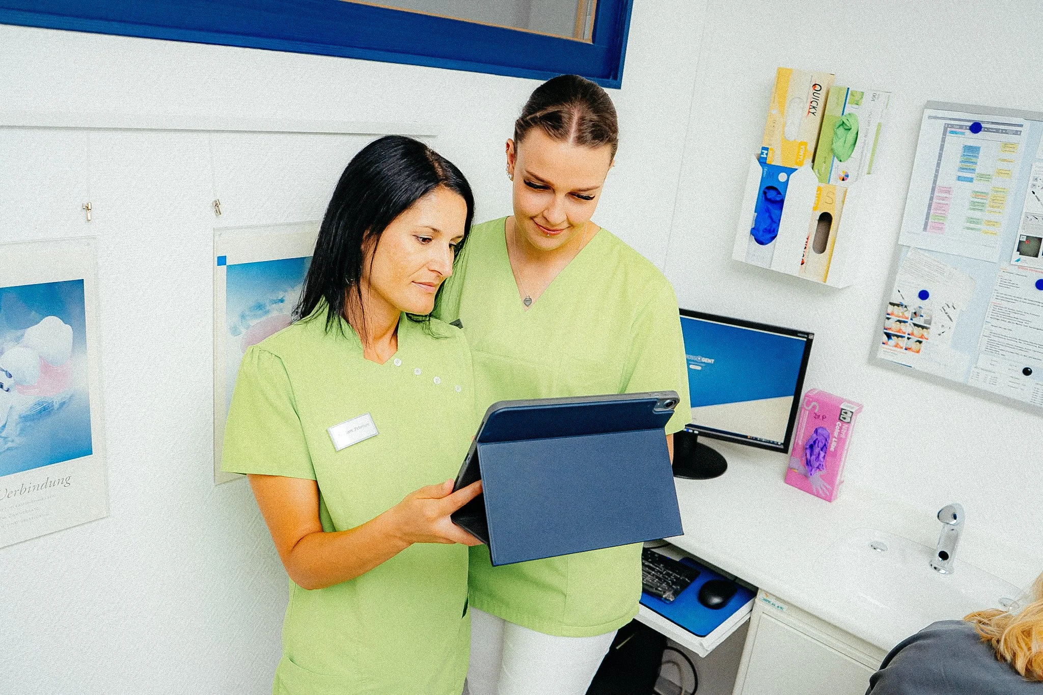 Zwei Pflegekräfte in grünem Uniform betrachten ein Tablet in einer Arztpraxis oder Klinik, im Hintergrund Arztutensilien und Poster an der Wand.