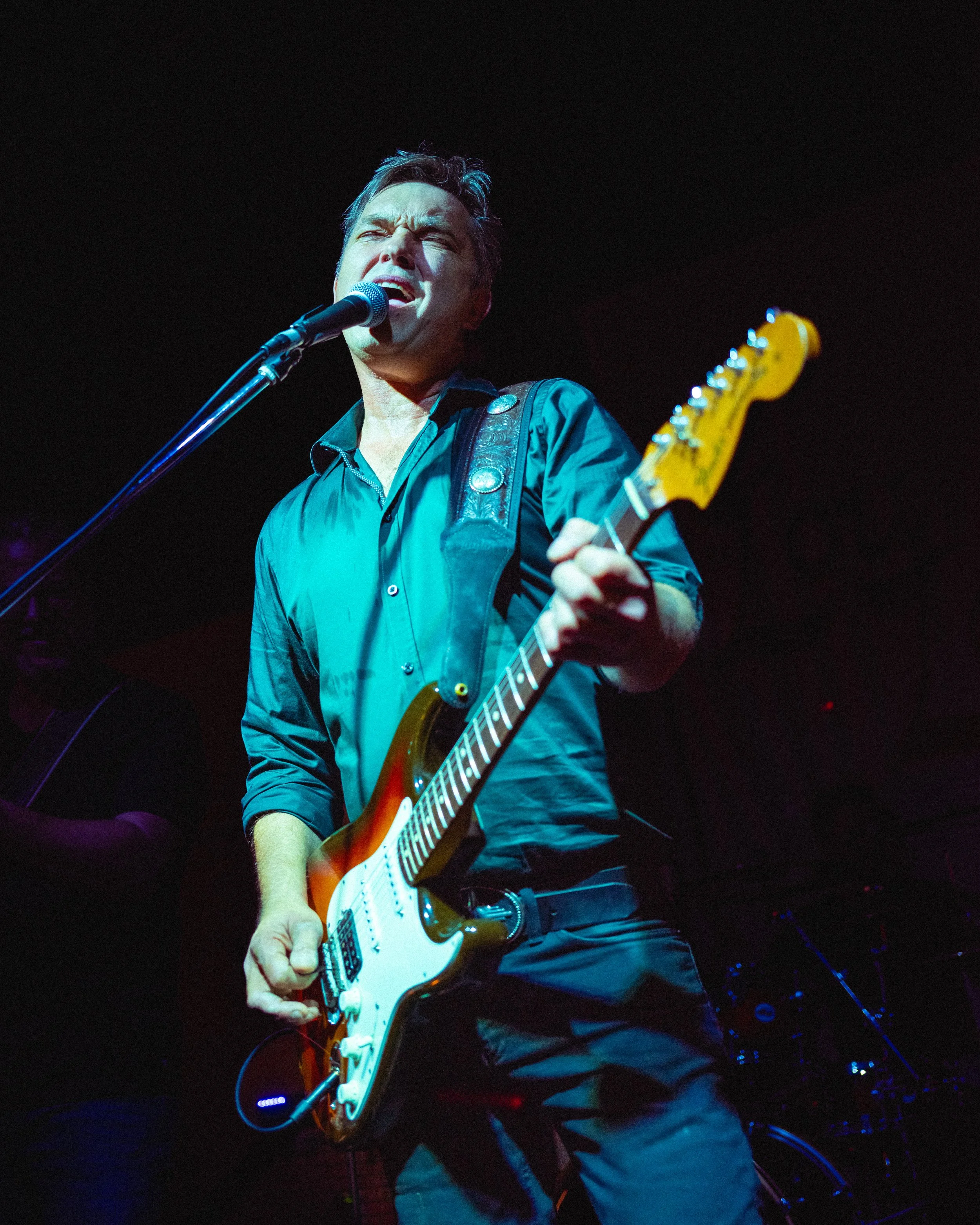 A man performing on stage with an electric guitar and singing into a microphone, wearing a dark shirt and black pants.