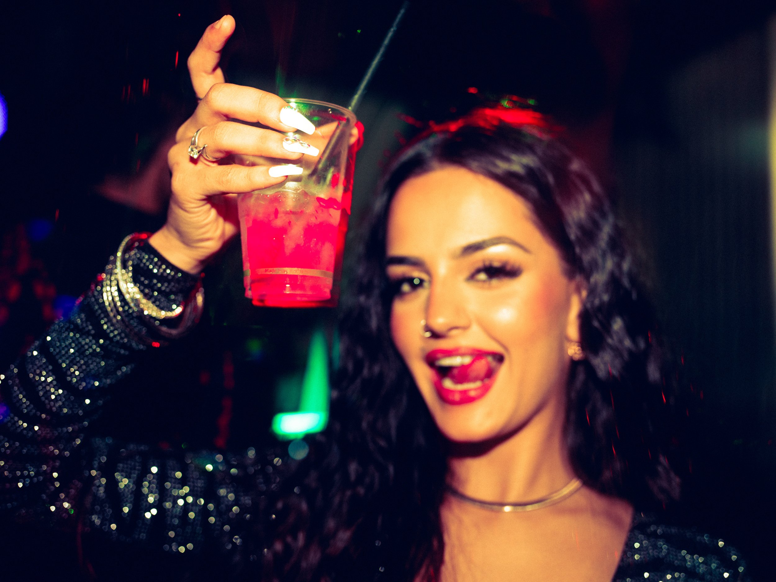 A woman with dark, curly hair and makeup, smiling and holding a pink drink with a stir stick in a nightclub with colorful lights.