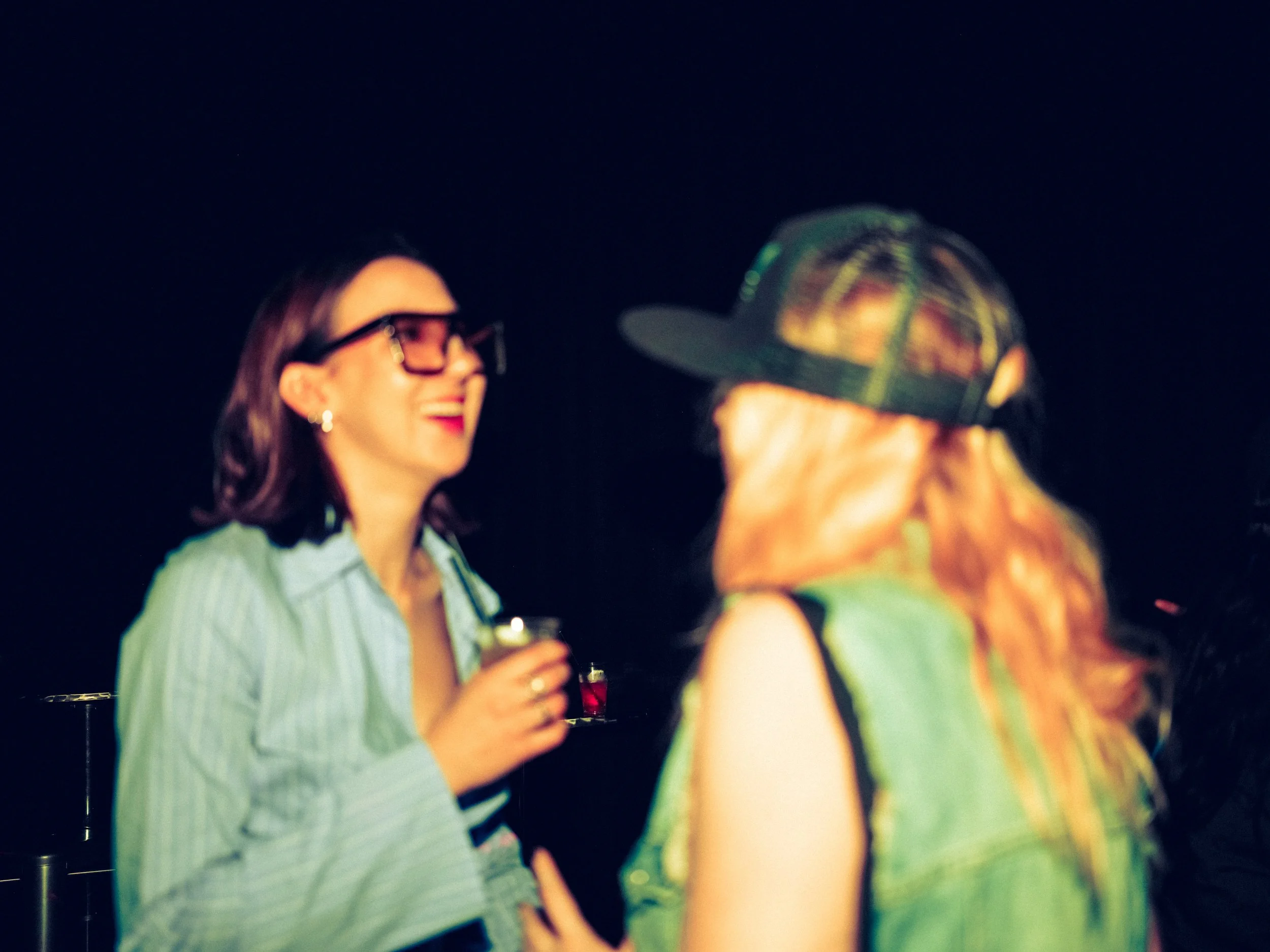 Two women conversing at night, one with shoulder-length dark hair, wearing large black glasses, red lipstick, a light blue shirt, and holding a drink, and the other with long blonde hair, wearing a black cap and a sleeveless vest, smiling.