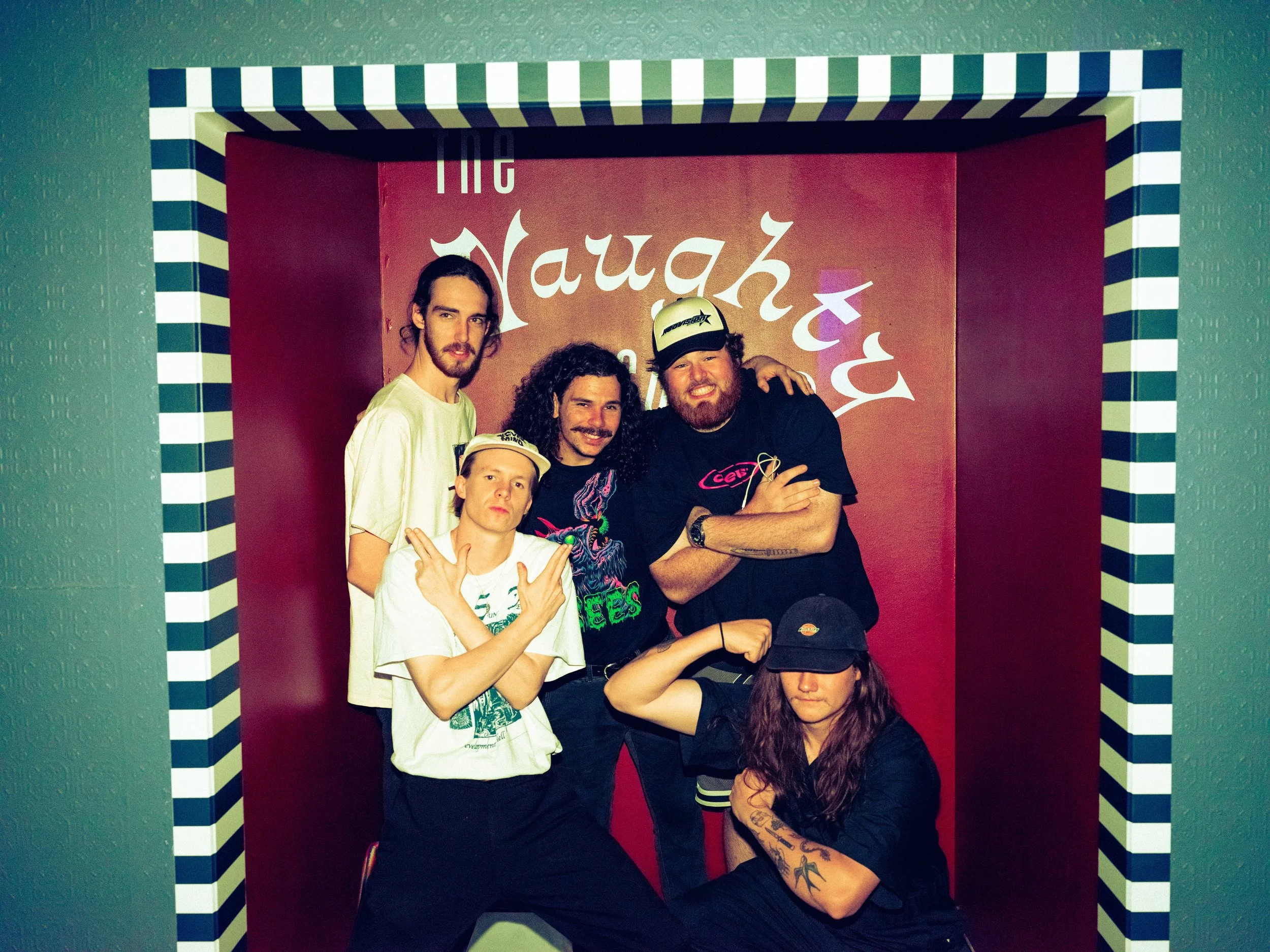 Group of six people posing in front of a red wall with white text that says 'The Laugh Factory.' Four men and two women, with some making gestures and wearing casual clothing and caps, smiling and standing close together.