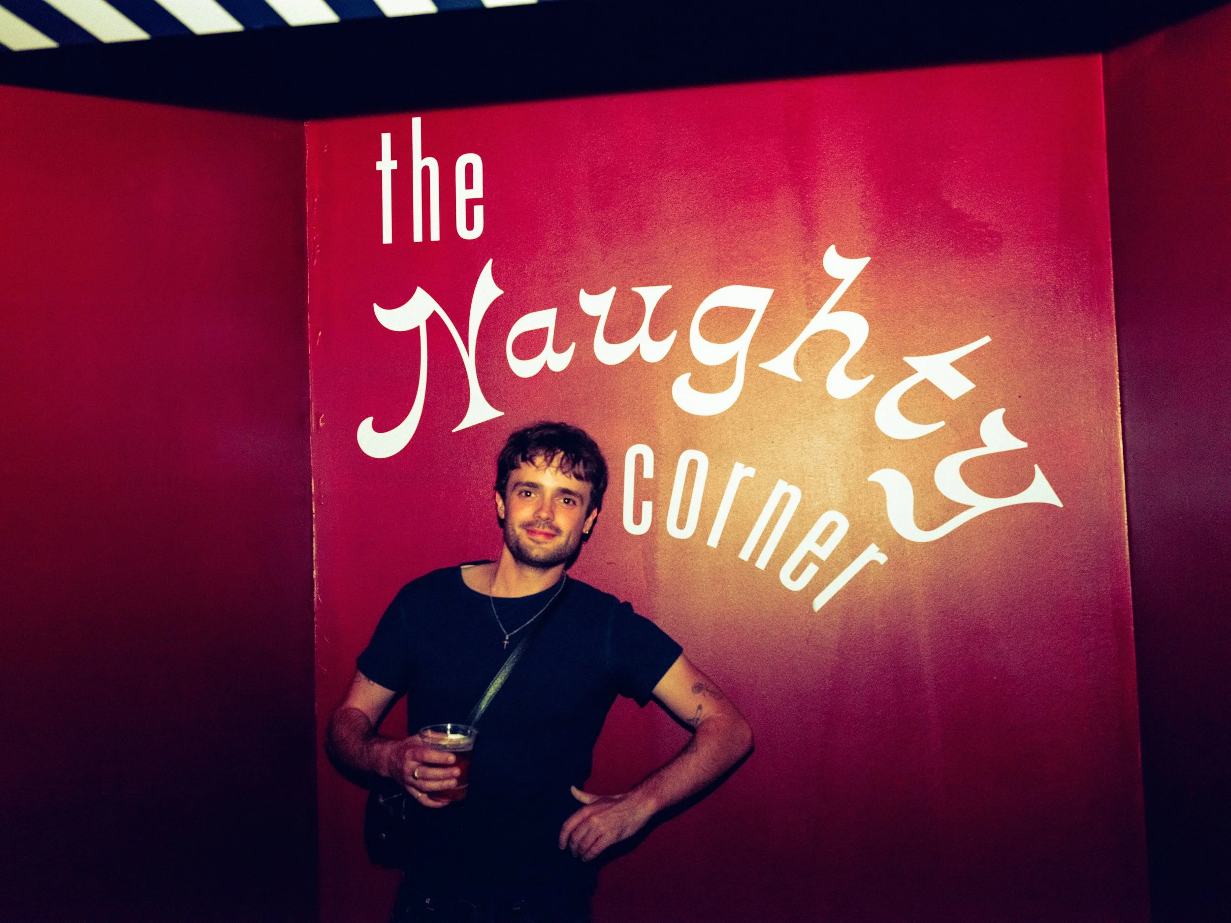 A young man with dark hair and a beard, wearing a black t-shirt, holding a drink, standing in front of a red wall with white text that reads 'the Naugh'ty corner.'