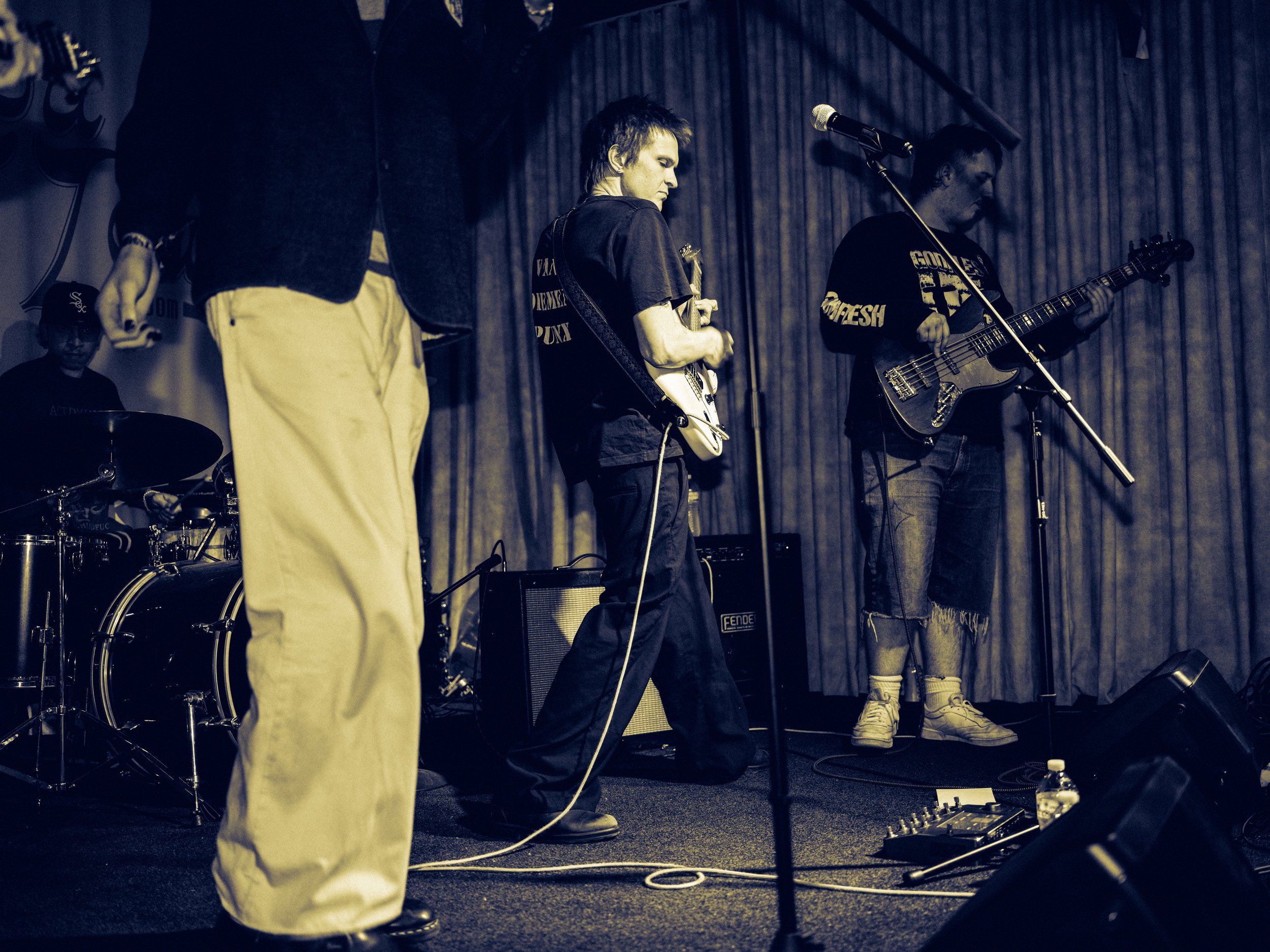 A band performing live on stage with multiple musicians playing guitars, drums, and vocals, under stage lighting with a curtain backdrop.
