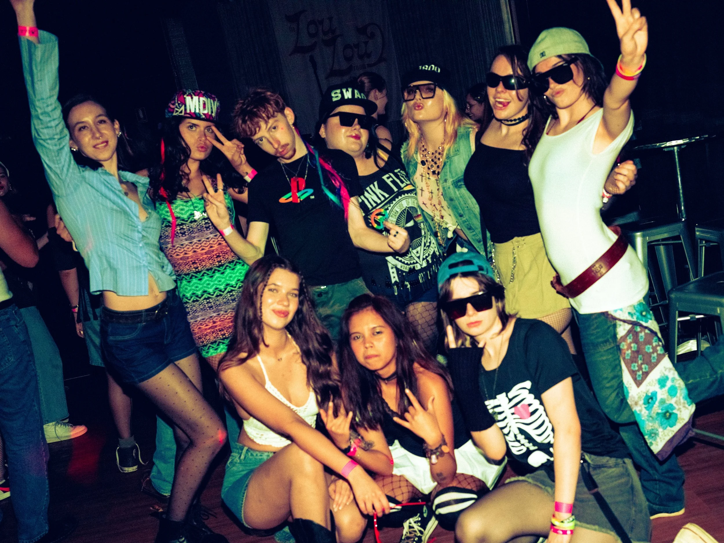 Group of young people at a party, wearing colorful outfits, sunglasses, and making peace signs or posing for the camera.