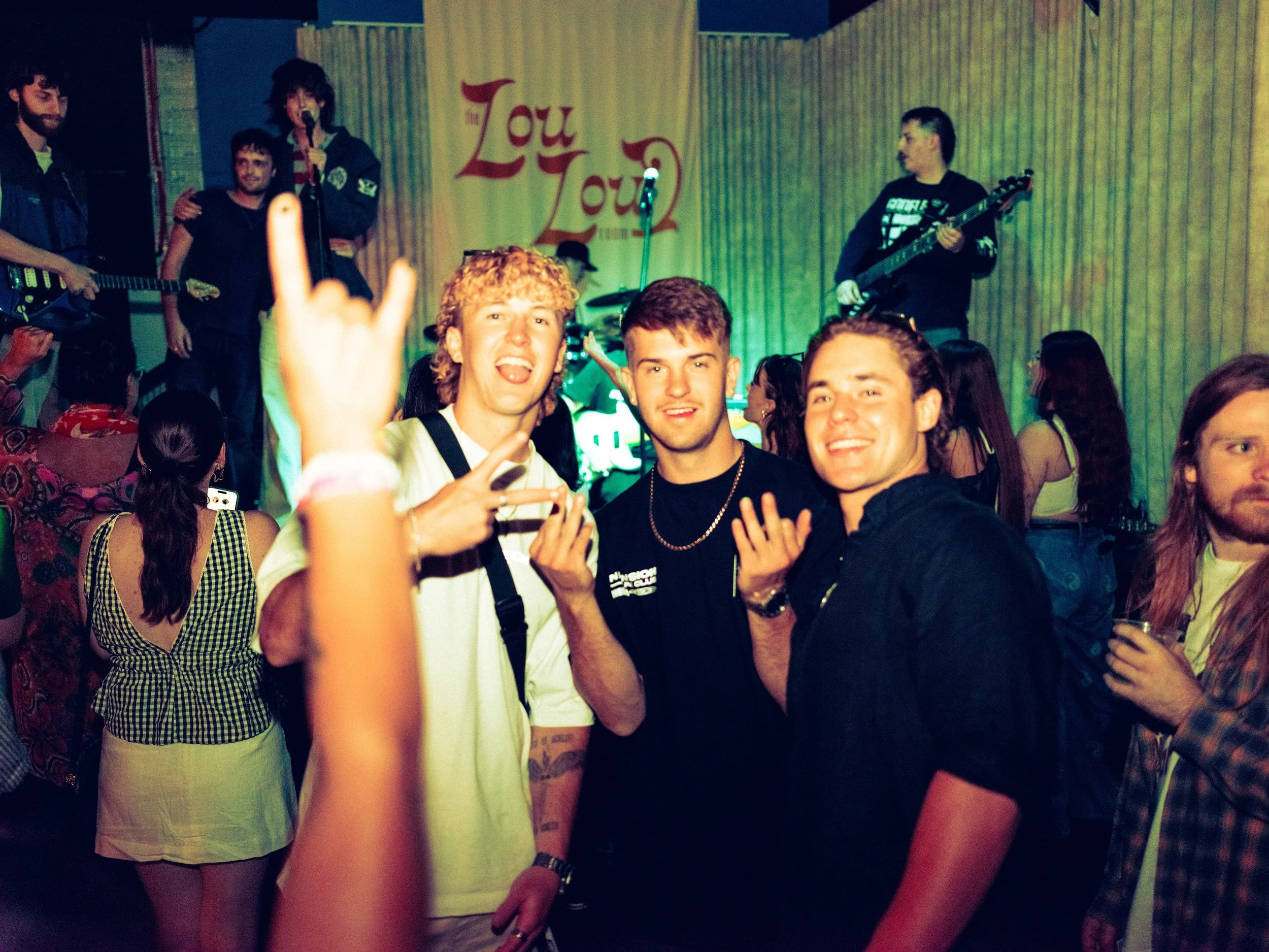 Group of young people enjoying a concert with a band on stage in the background, some making peace signs and smiling for the camera.