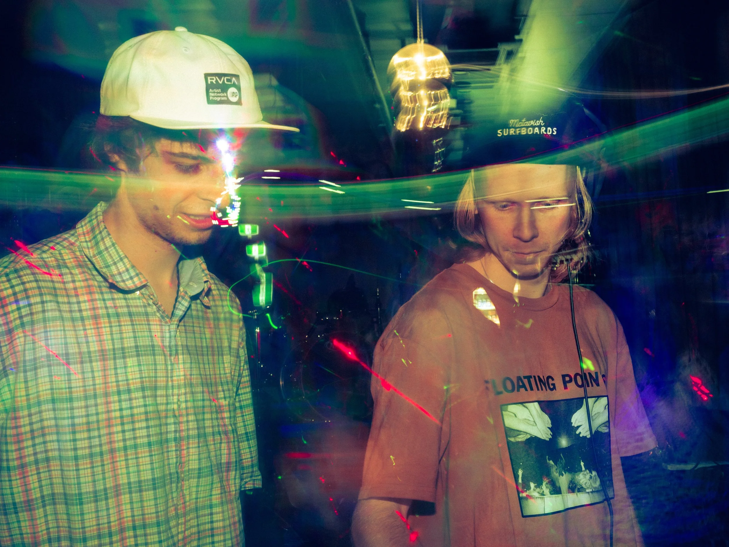 Two young men with light skin in a colorful, dimly lit nightclub or party scene, one wearing a white cap and plaid shirt, the other wearing a dark cap and a graphic t-shirt, surrounded by colorful laser light streaks.