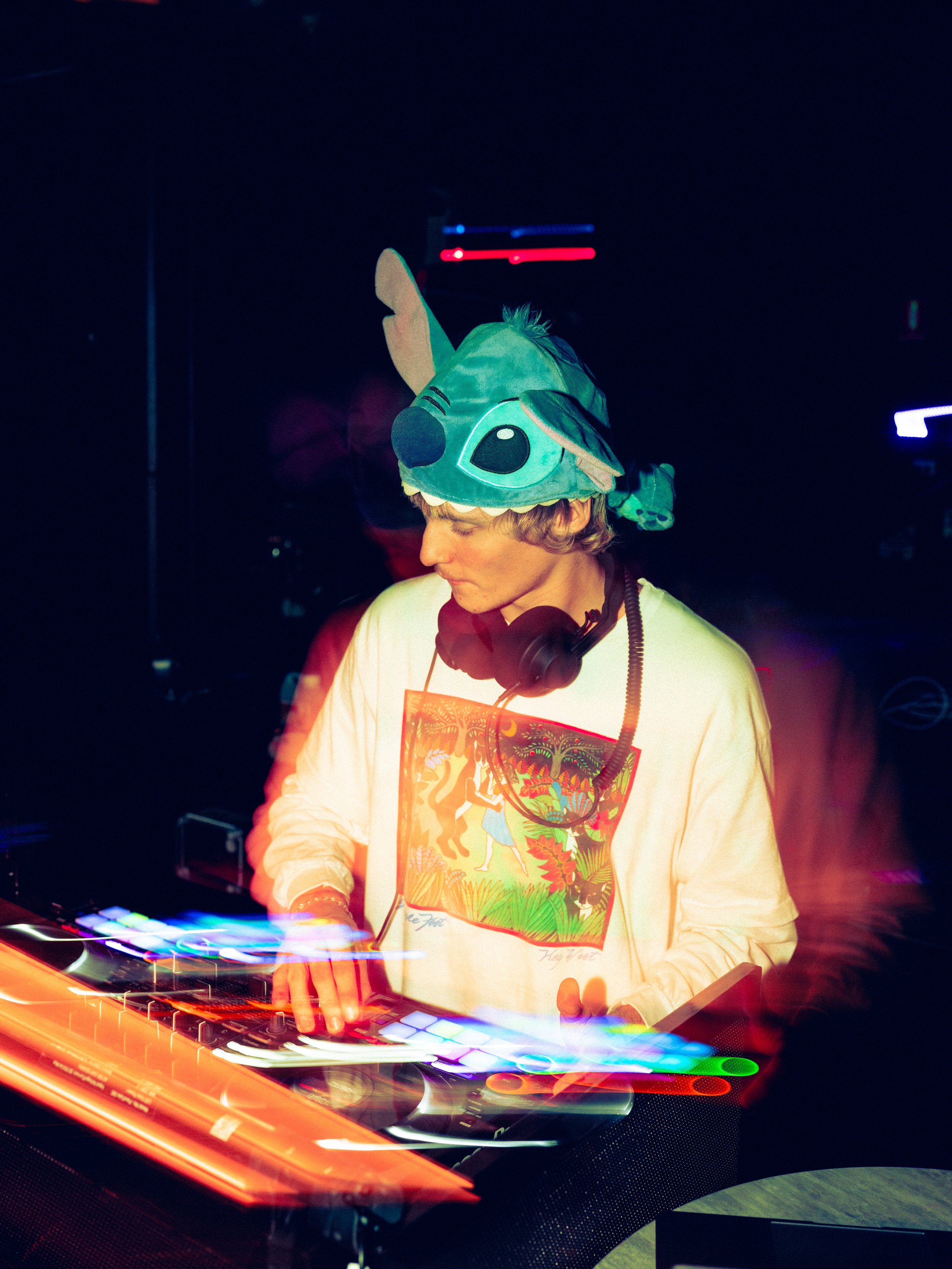 A DJ wearing a colorful Stitch hat, with headphones around their neck, playing music on DJ equipment in a dark club with vibrant lights.