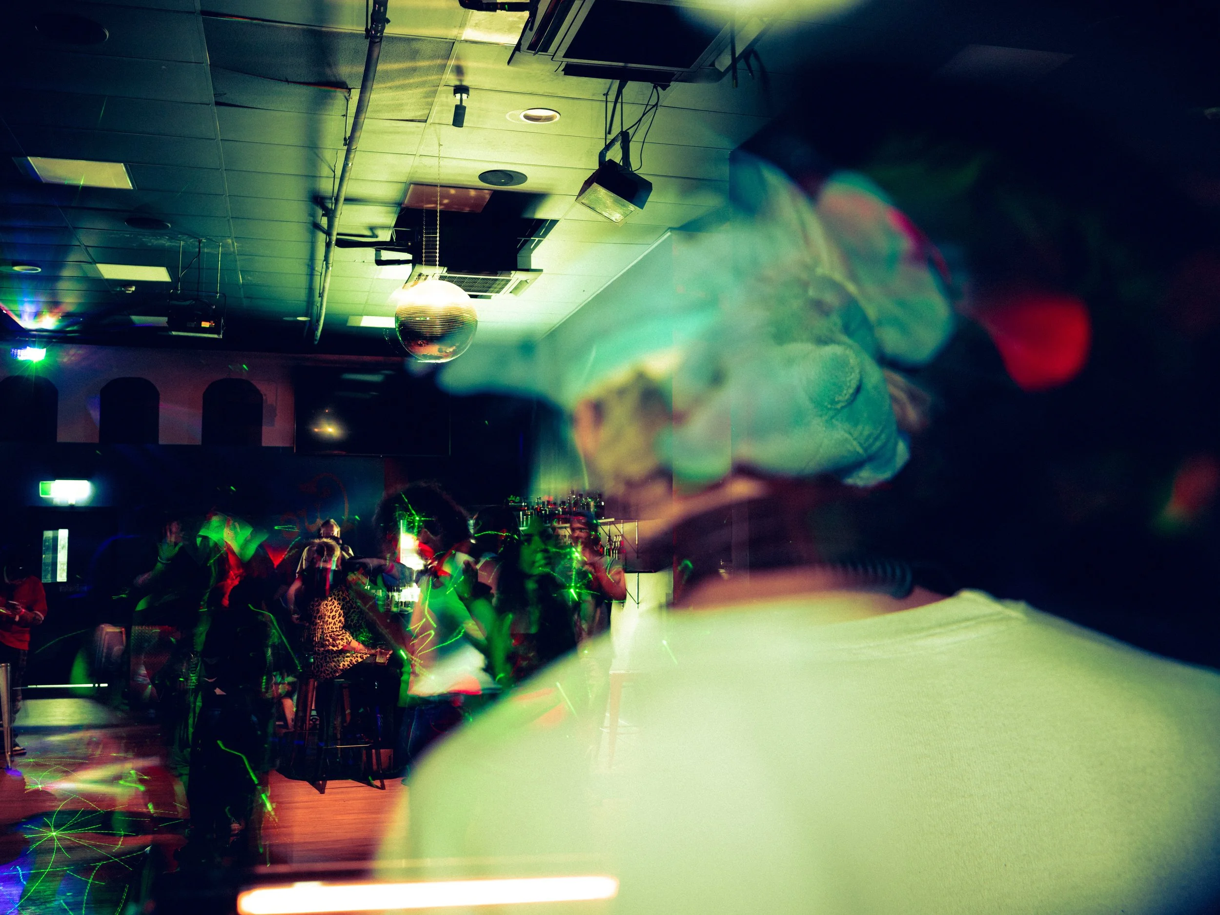 A person with multi-colored lights shining on their face, with a group of people dancing and socializing in a dimly lit room with a disco ball overhead.