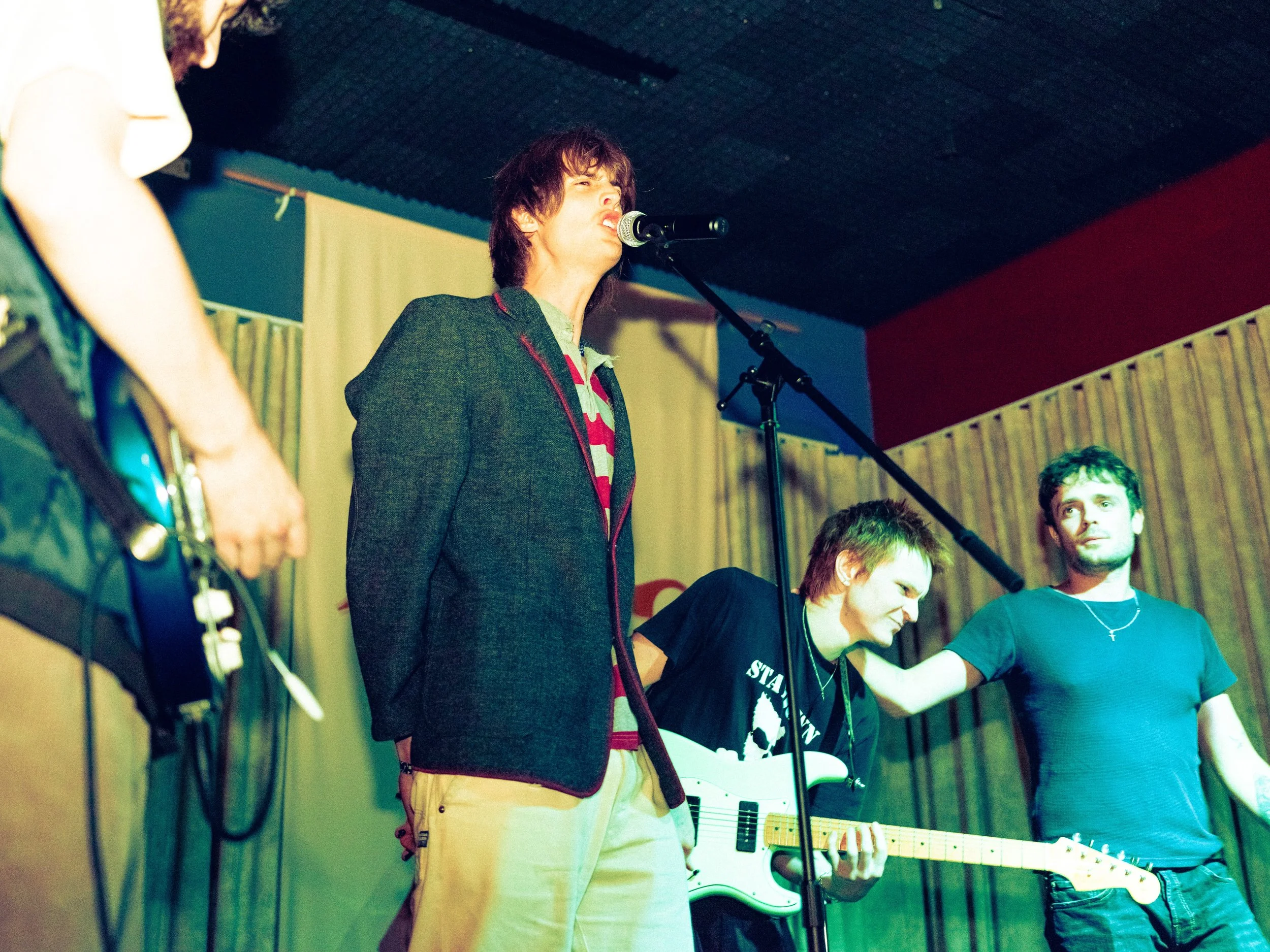 A band performing on stage with four members visible. The lead singer, in a blazer, is singing into a microphone. A guitarist in a black t-shirt is standing next to him, and another musician in a blue t-shirt is also on stage.