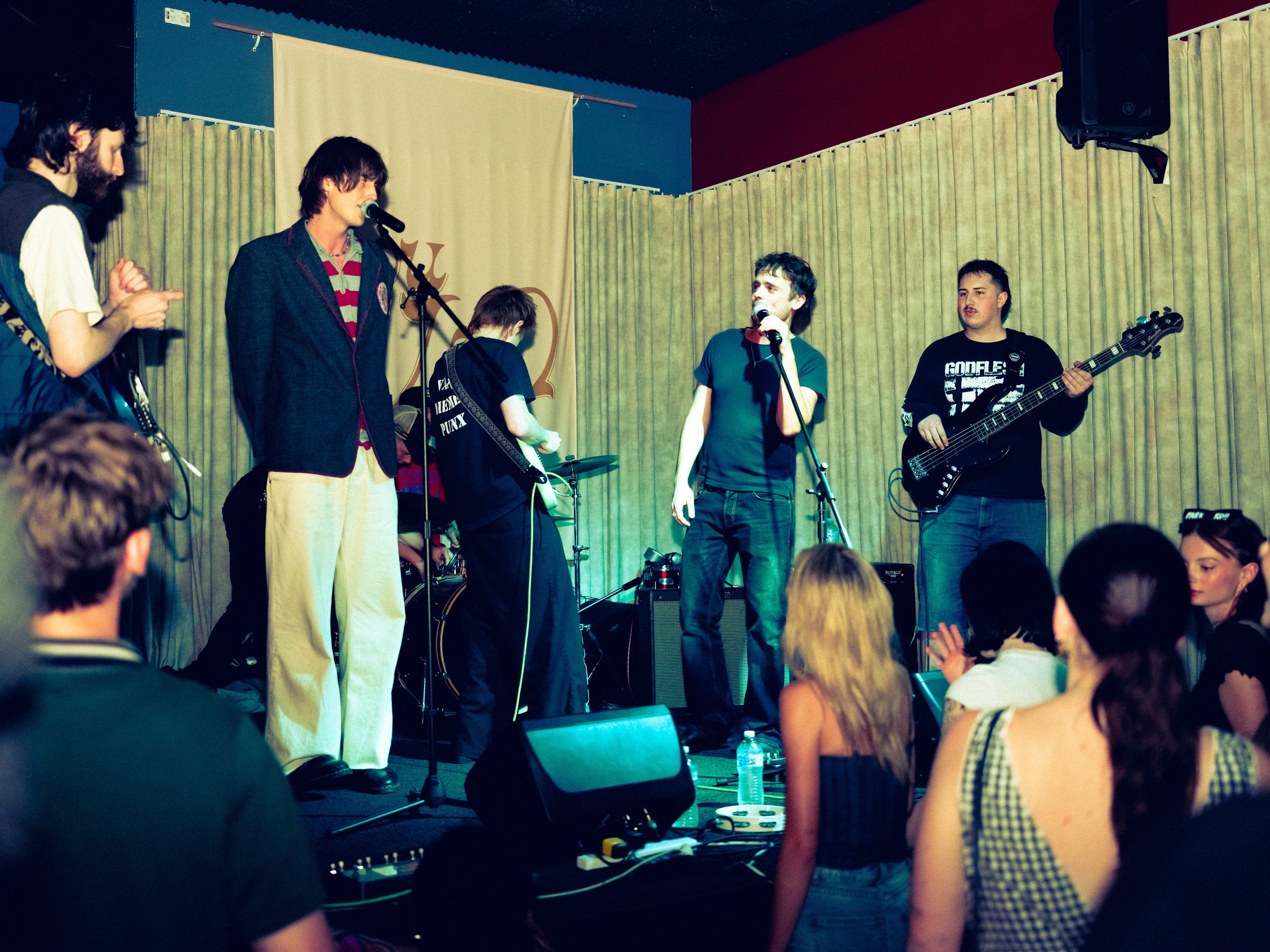 A band performs on stage at a music venue, with four members playing instruments and singing, while an audience watches.