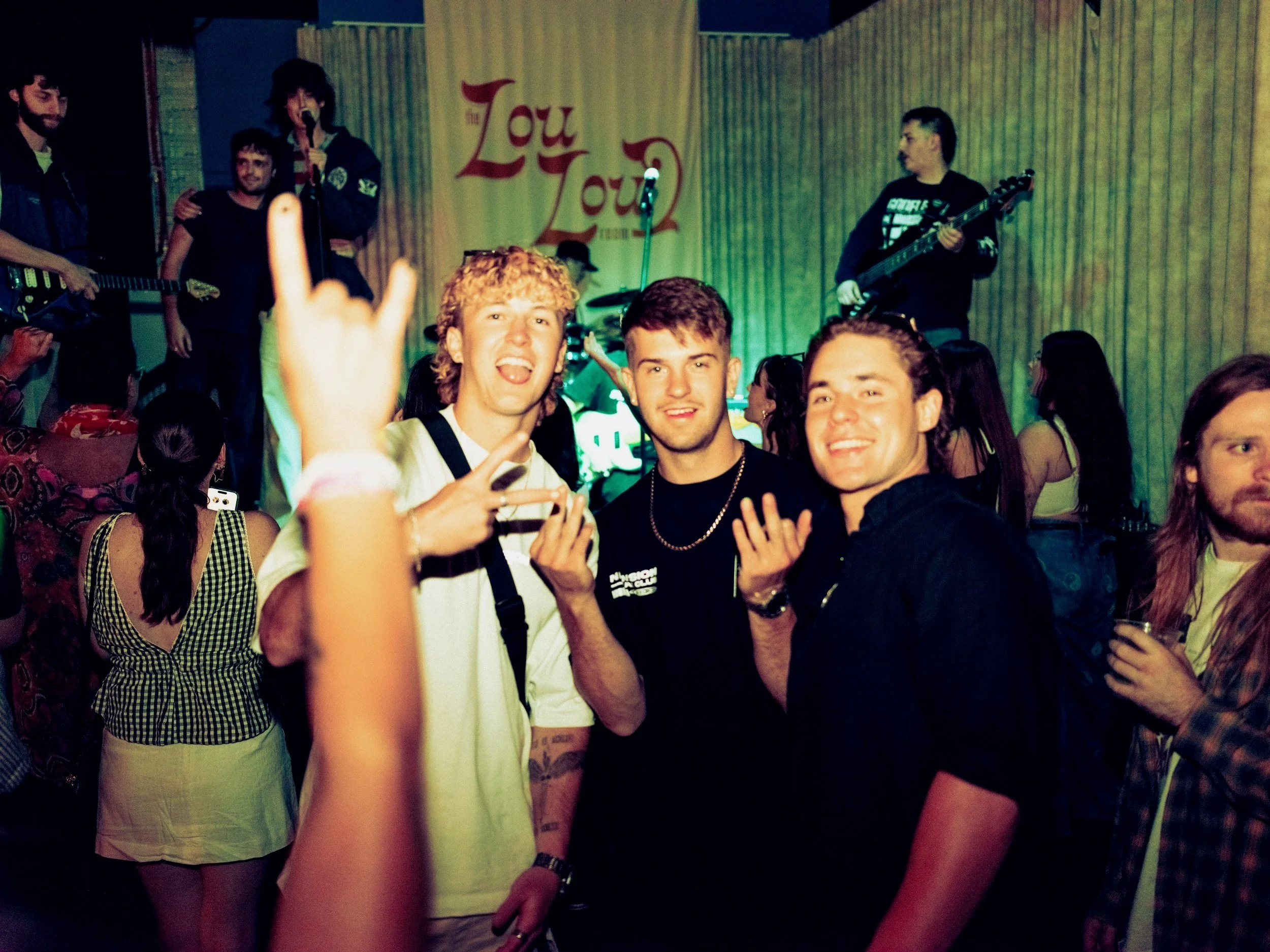 Group of young people at a concert or party, with a band performing on stage in the background.