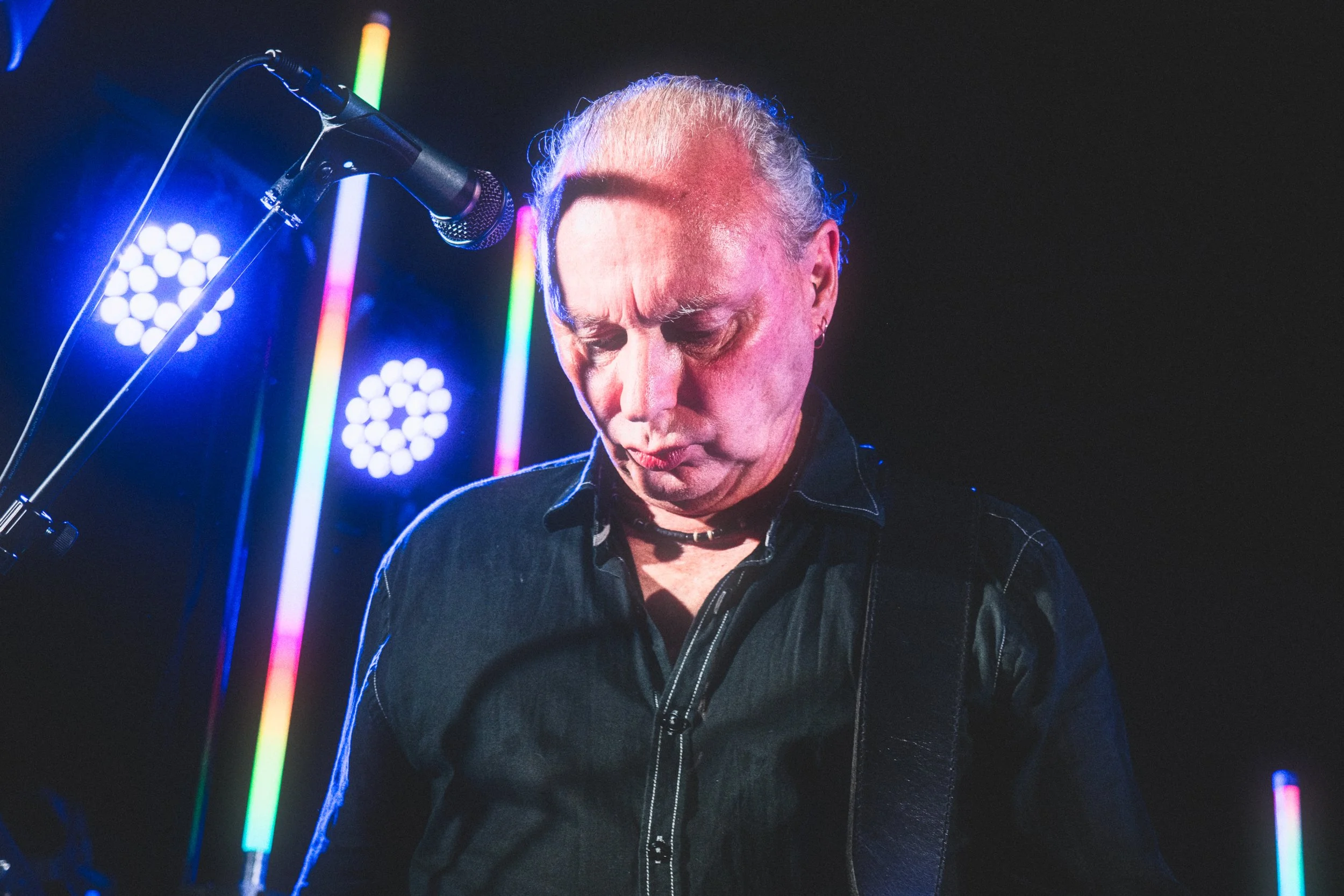 A man with short, curly, gray hair playing guitar or bass, with his eyes closed, amidst colorful vertical neon lights and stage lights.