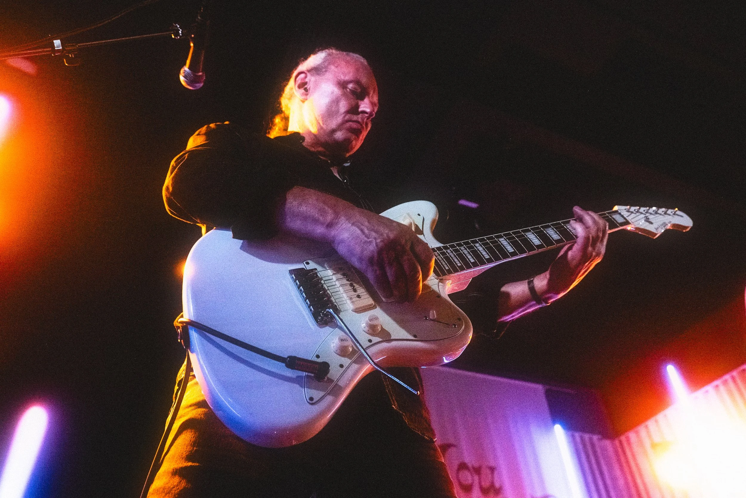 A guitarist playing an electric guitar on stage with colorful lights behind him.