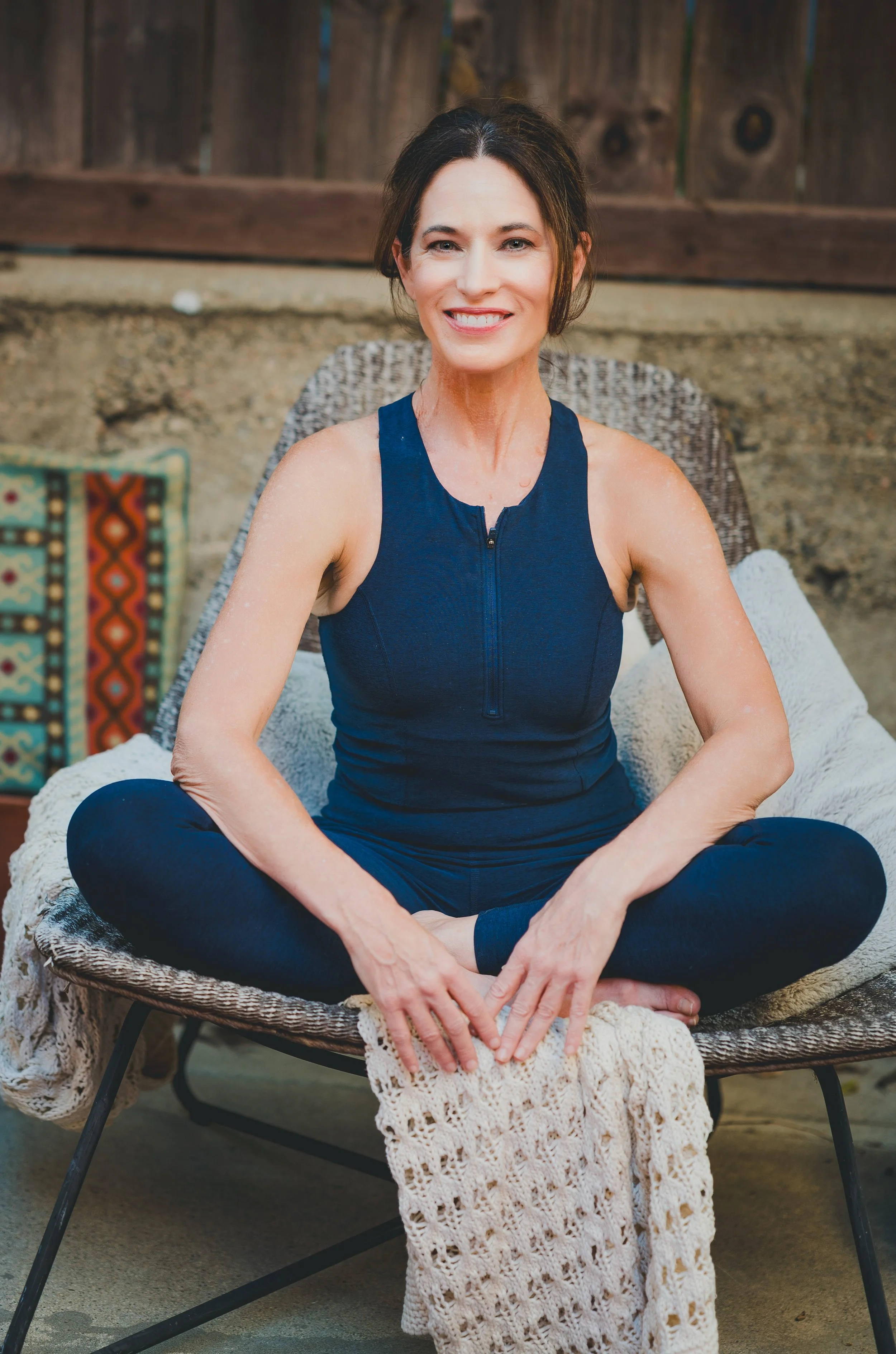 Woman with dark brown hair smiling and sitting cross-legged on a woven chair outdoors, wearing a sleeveless navy blue athletic top.