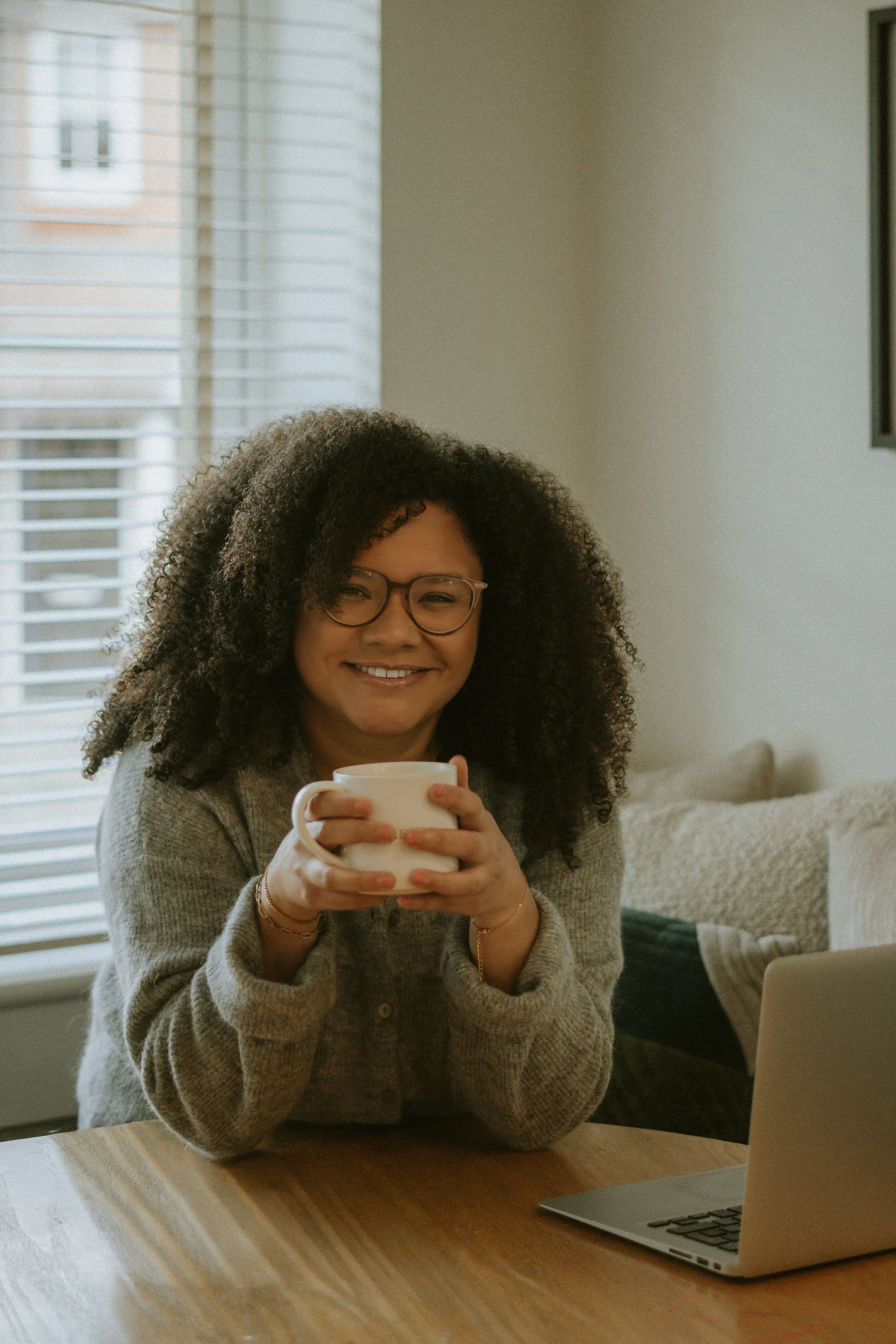 woman sat at table, holding a mug of drink with a laptop in front of her on the table