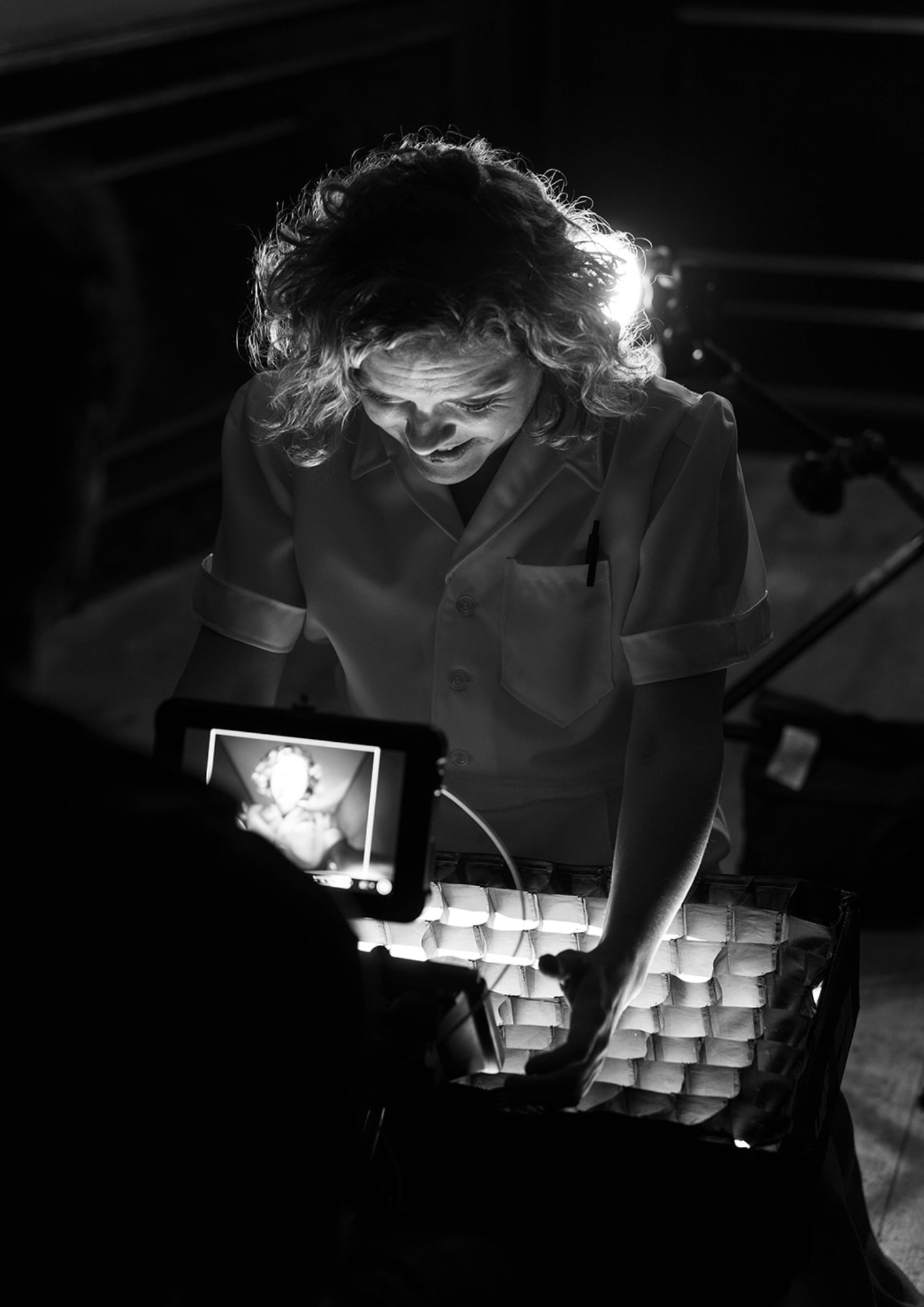 Black and white photo of a woman with curly hair looking down at a lit object with a curious expression, with a recording device capturing her in the foreground.