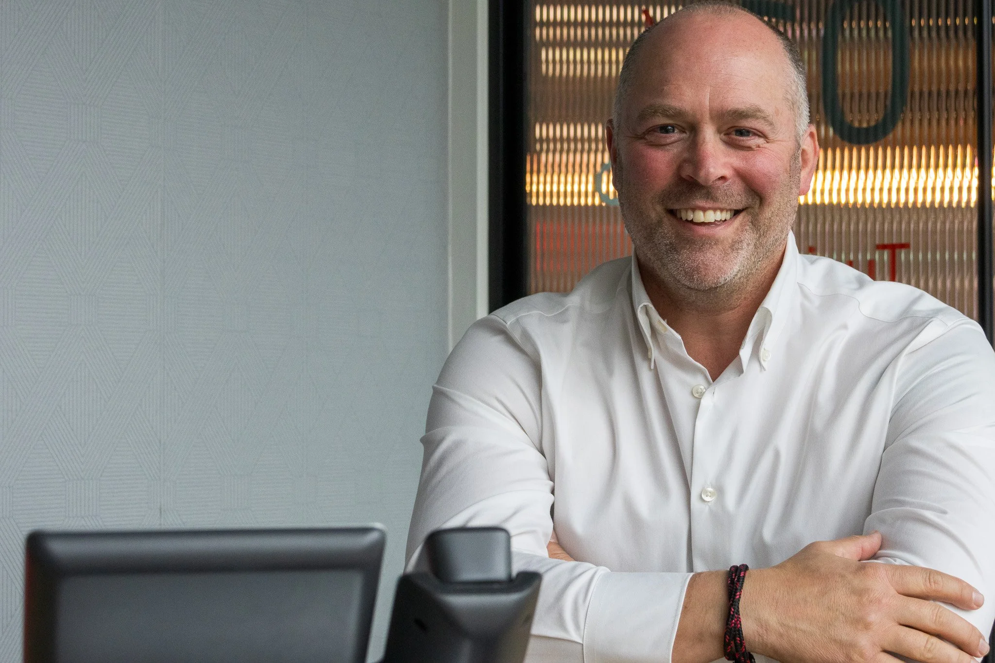 A smiling man in a white shirt sitting at a desk with a phone and a laptop in an office setting.
