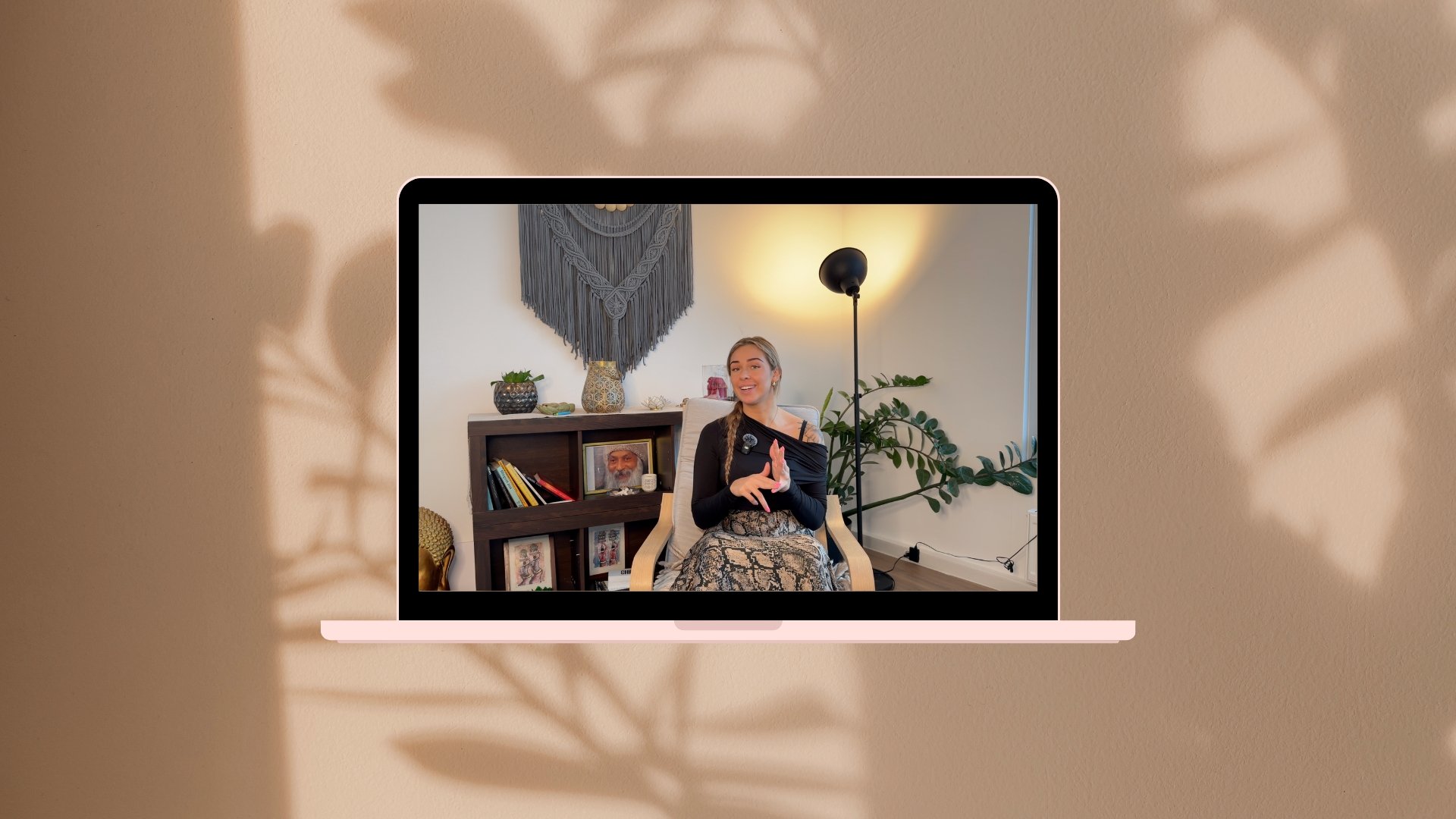 A woman sitting in a room, smiling and gesturing with her hands, visible on a computer screen. The room has decorative plants, framed photographs, and a large fan-shaped wall hanging.
