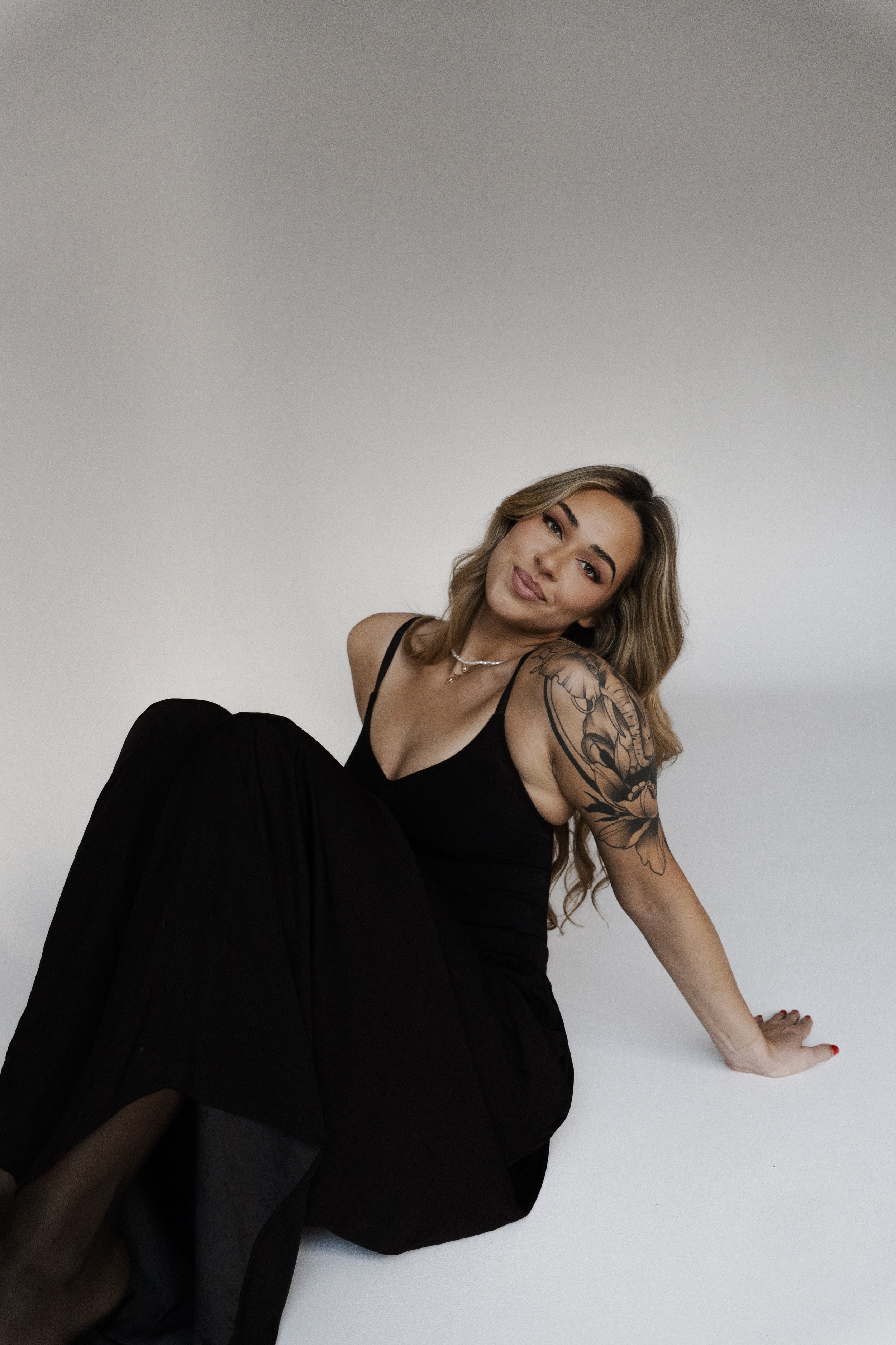 A woman with long wavy hair wearing a black dress, sitting on the floor against a plain white background, looking at the camera with a slight smile, showing a detailed tattoo on her upper arm.