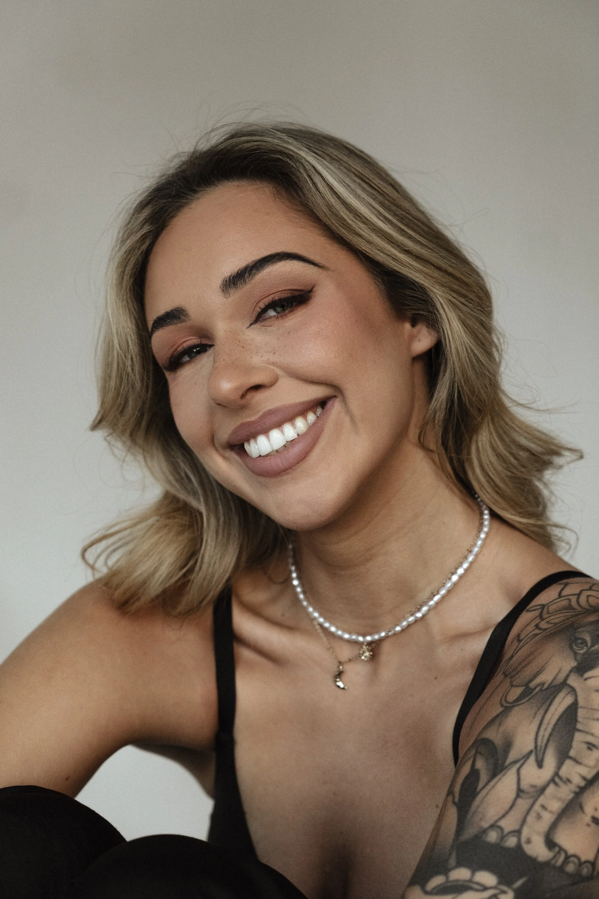 A smiling young woman with blond hair styled in loose waves, wearing makeup including winged eyeliner and nude lipstick, a pearl necklace with a moon and star pendant, a black tank top, and a detailed tattoo on her left arm, against a plain white background.