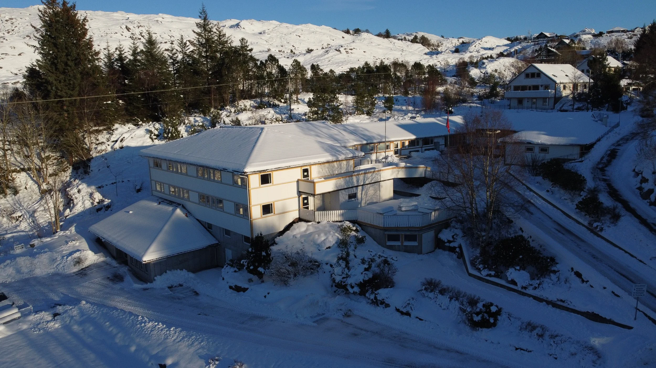 Et hvitt hus på fjellet, dekket av snø, med flere plan, store vinduer og rundt terrasse, omgitt av snø og trær.
